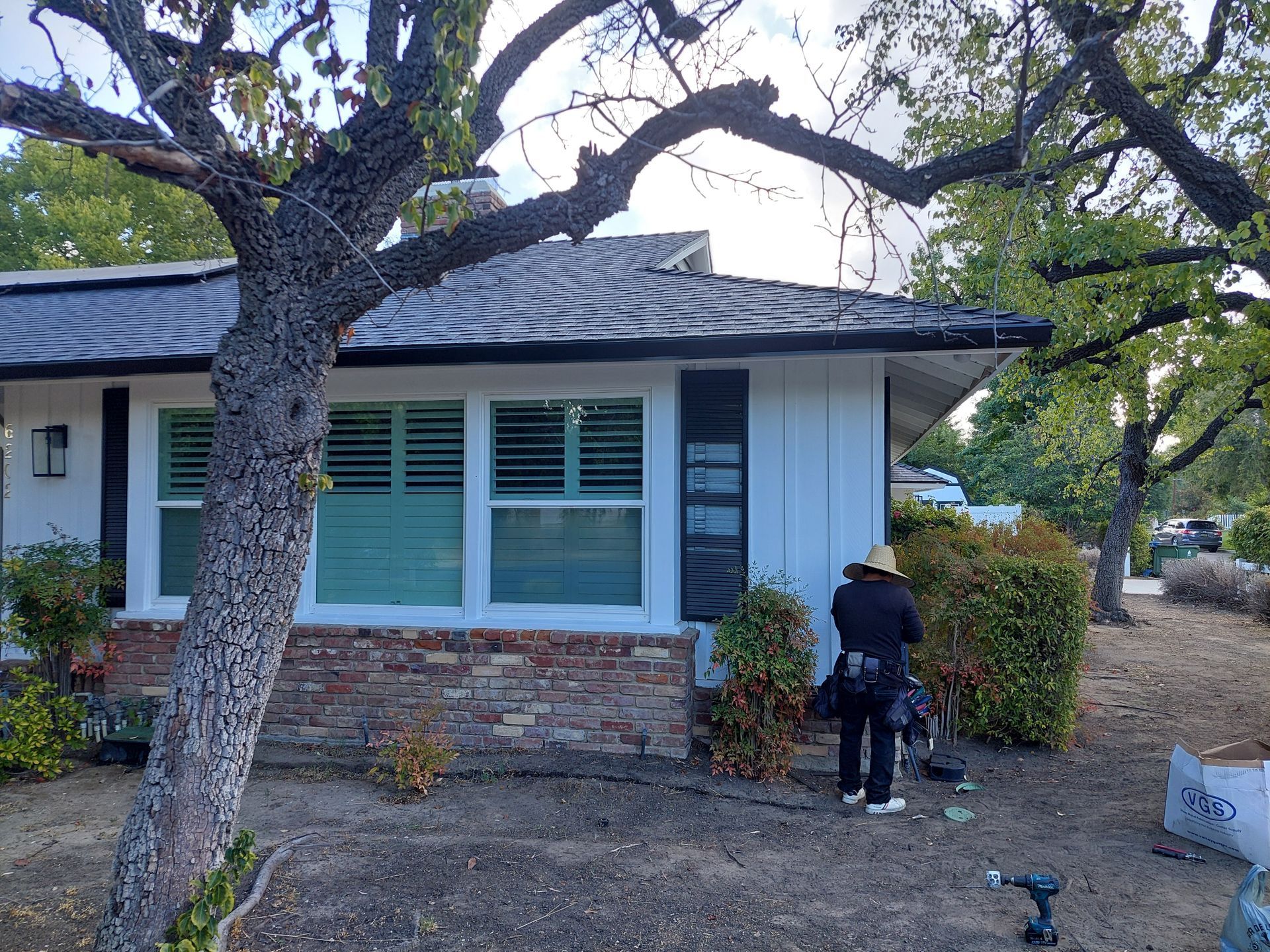 Person working near a house with blue shutters and a stone facade under large tree branches