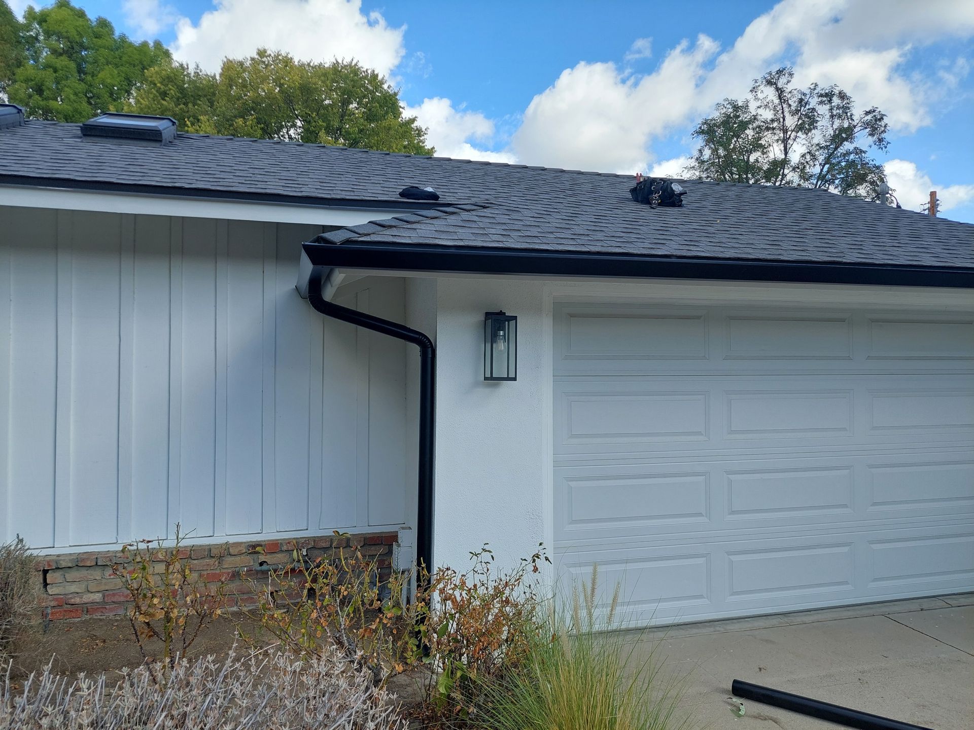 House exterior with white siding, black gutter, garage door, and overgrown front shrubs