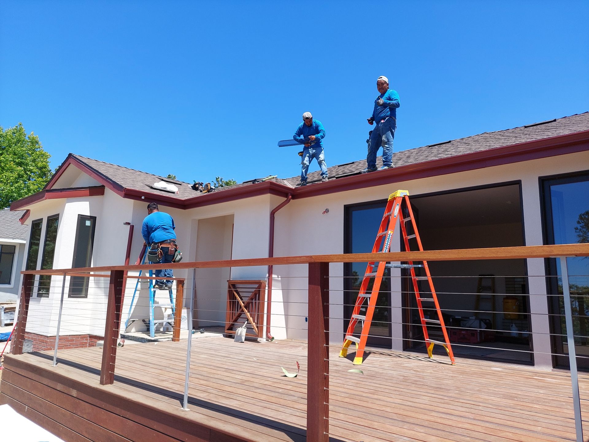 Workers on a rooftop repairing a house under a clear blue sky, with ladders and scaffolding in front.