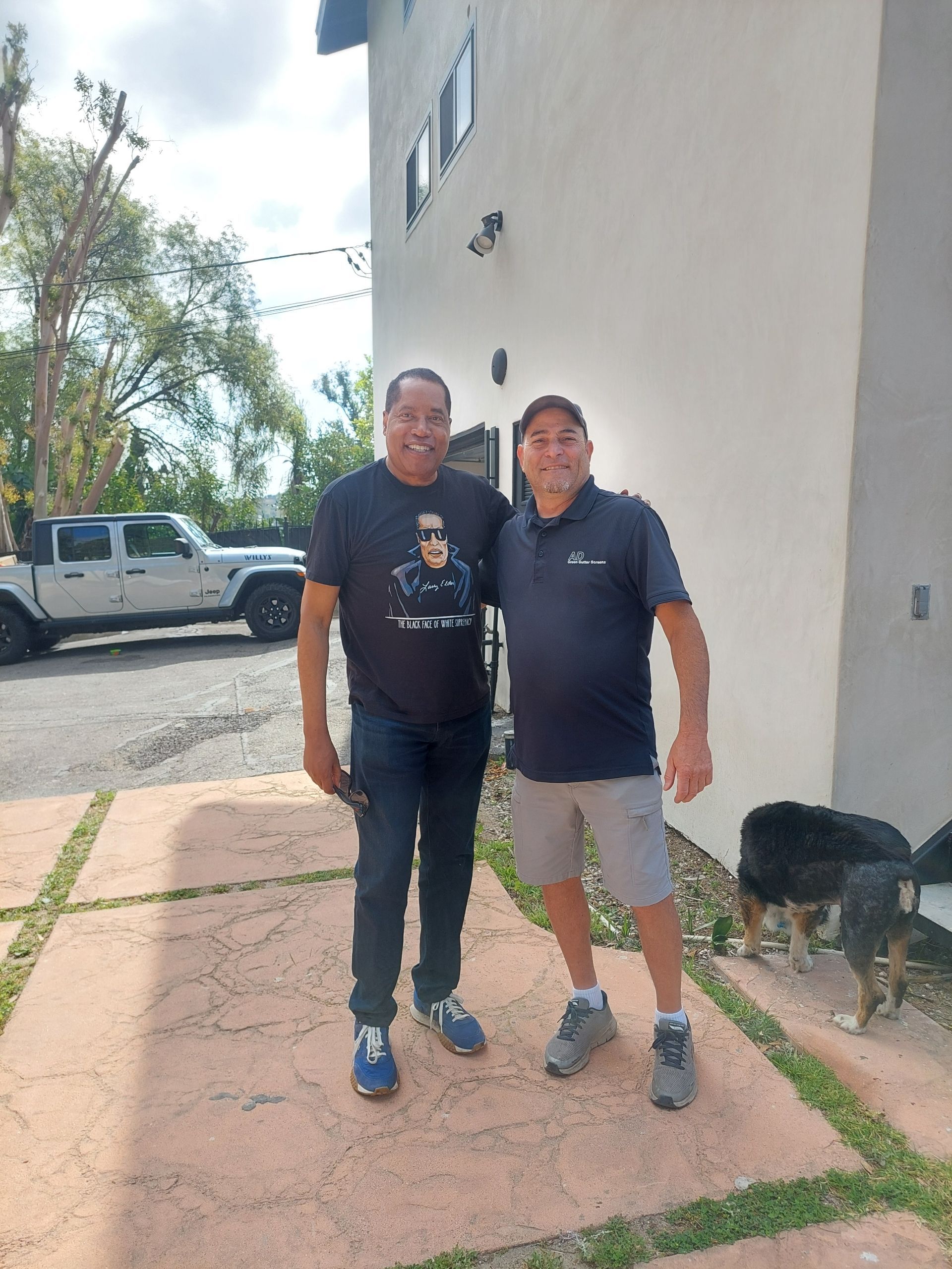 Two people standing together outdoors beside a building and parked vehicles, with a dog nearby.