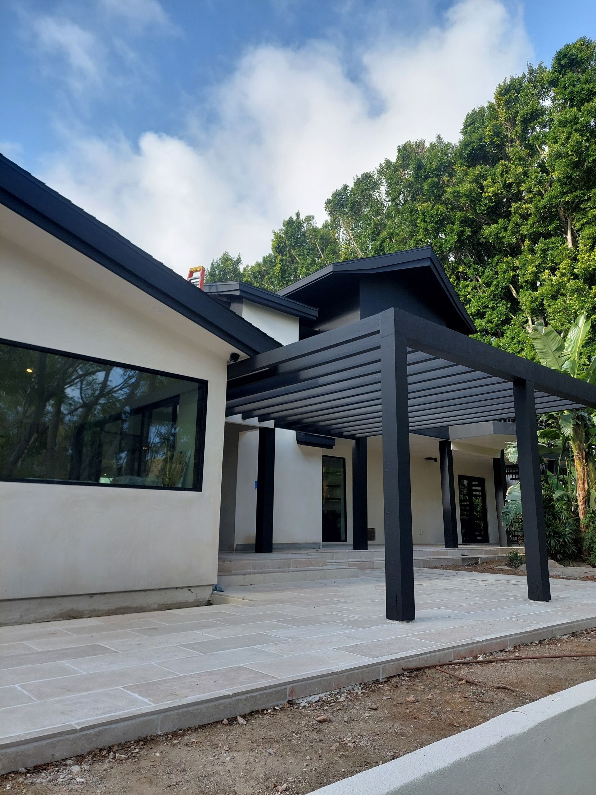 Modern house exterior with black pergola, white walls, and stone patio under trees