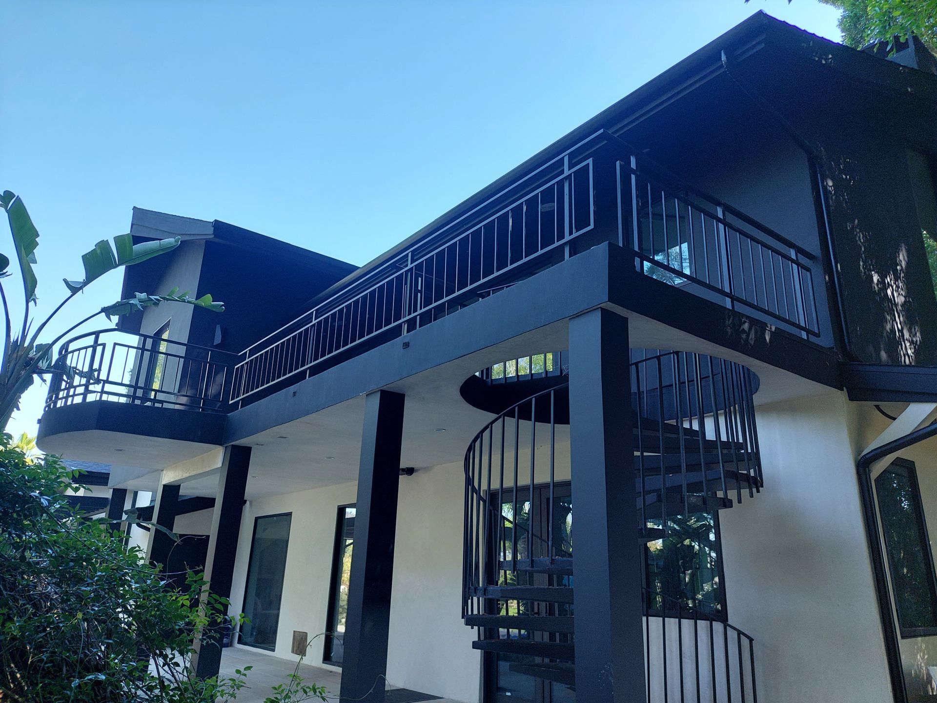Two-story modern house with white walls, dark balcony railings, and a sloped roof in a lush tropical setting