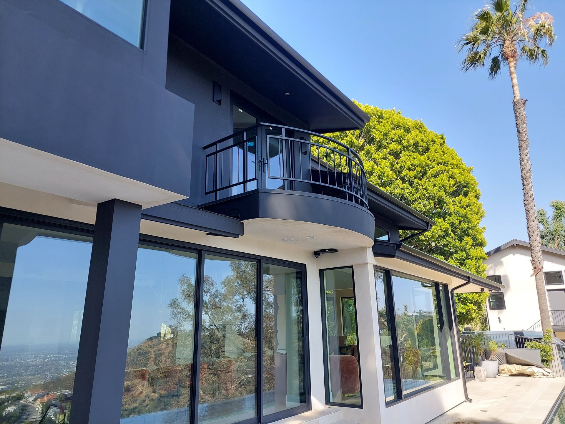 Modern blue house exterior with large glass windows, balcony, and palm trees in bright sunlight