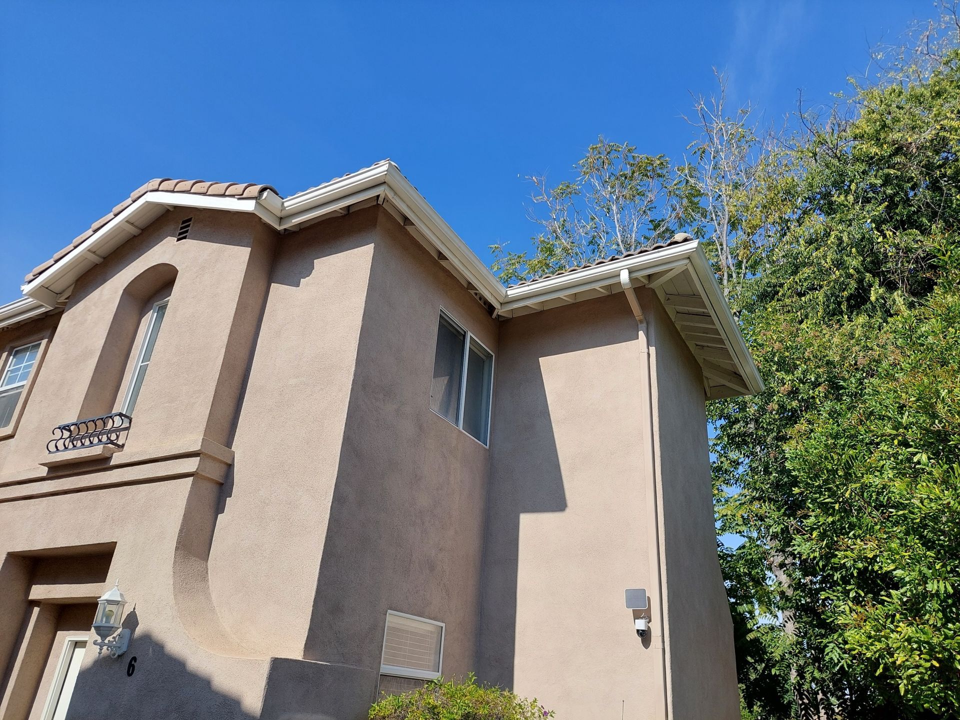 Two-story beige stucco house viewed from below against a blue sky and trees