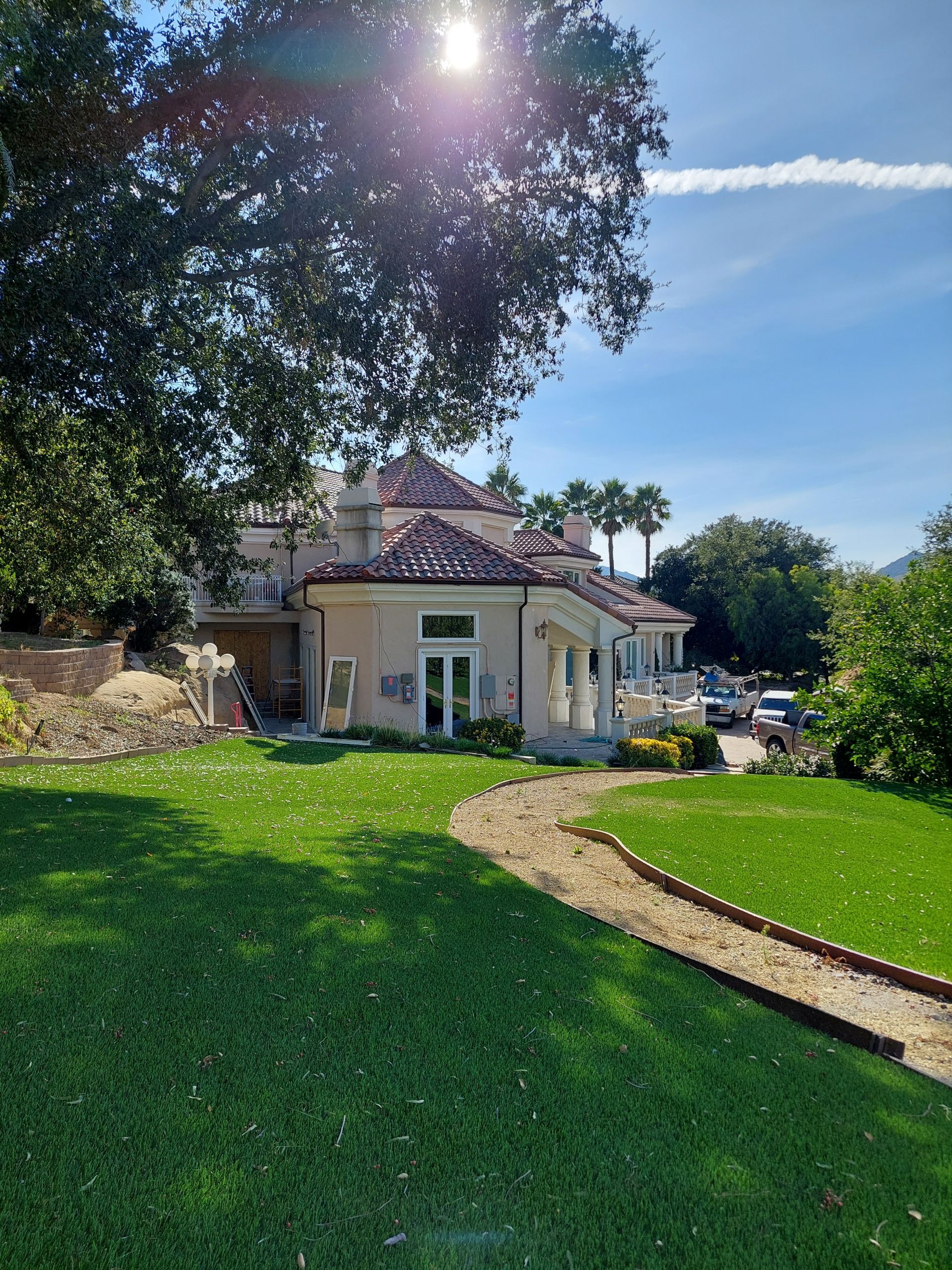 Sunlit beige house with red-tiled roof and green lawn, viewed along a curved stone path