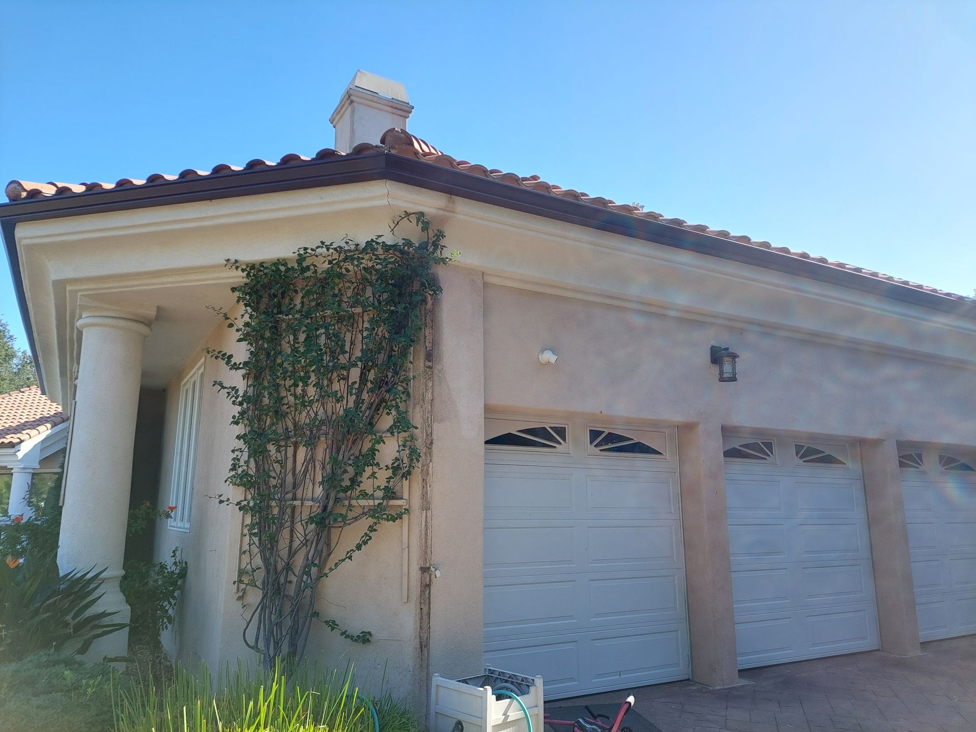 Single-story stucco house with white garage doors, tiled roof, and climbing vines on the side wall