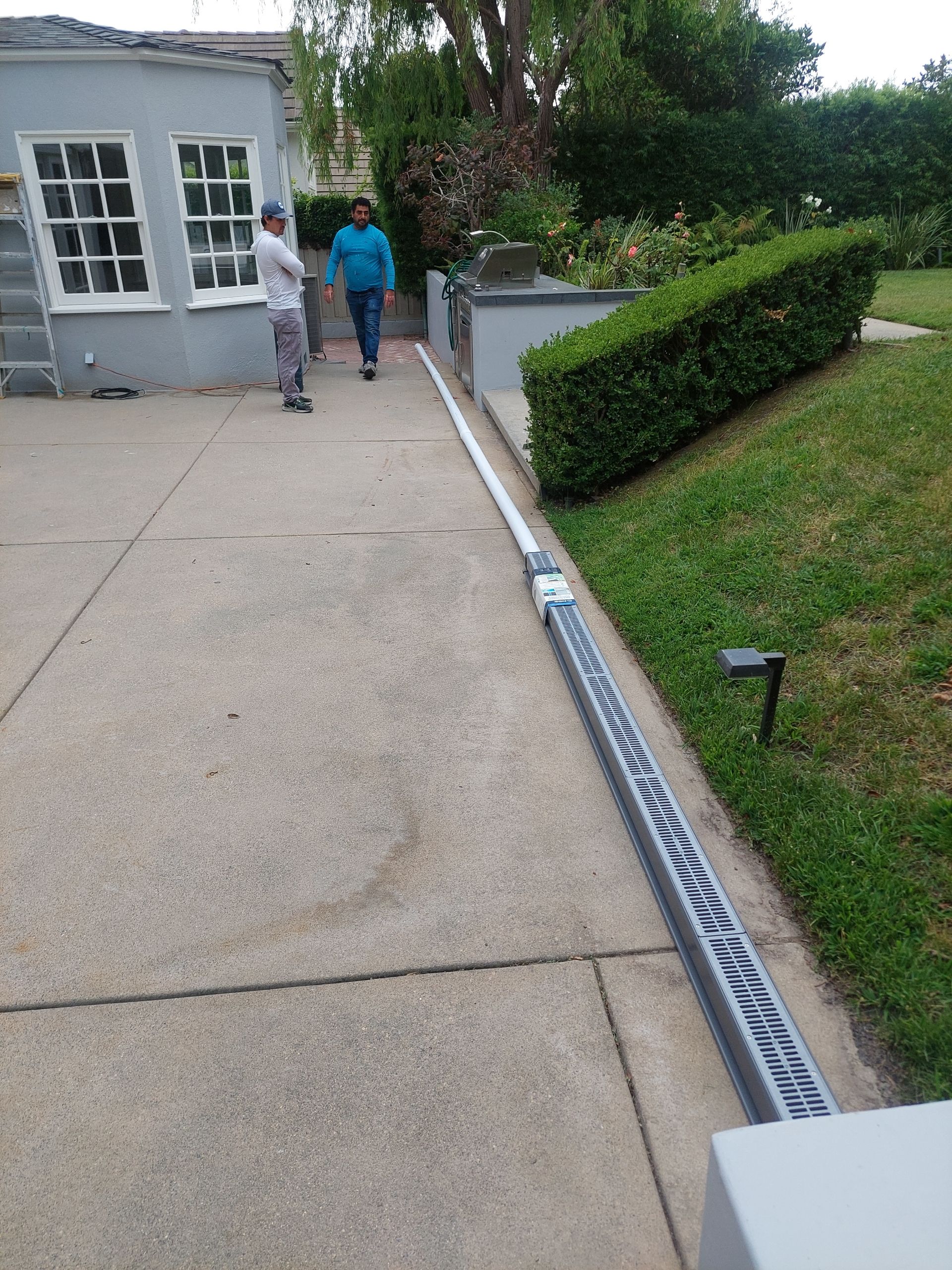 Two people walking beside a house, next to a long downspout and trimmed hedge.