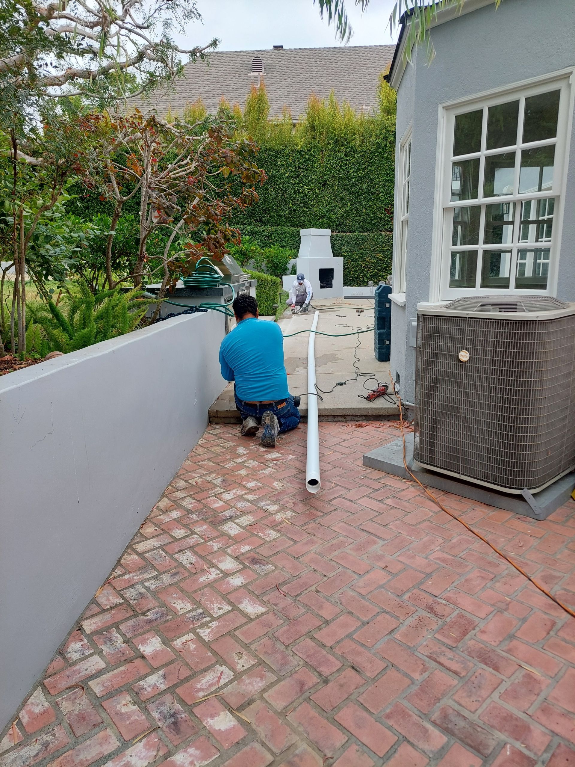 Person kneeling on a brick patio beside a white wall, working near a pool pump and air conditioner unit.