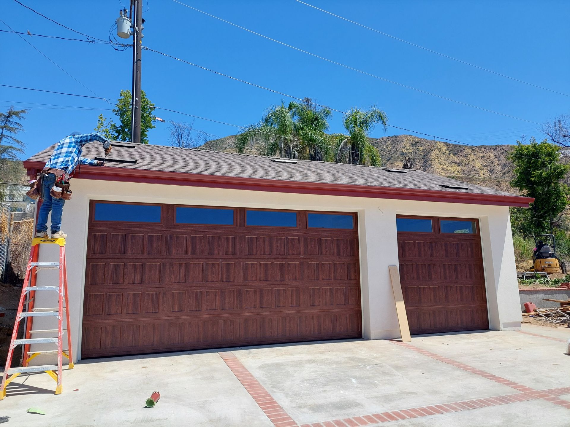 Worker on ladder repairing roof of white garage with brown doors under a bright blue sky