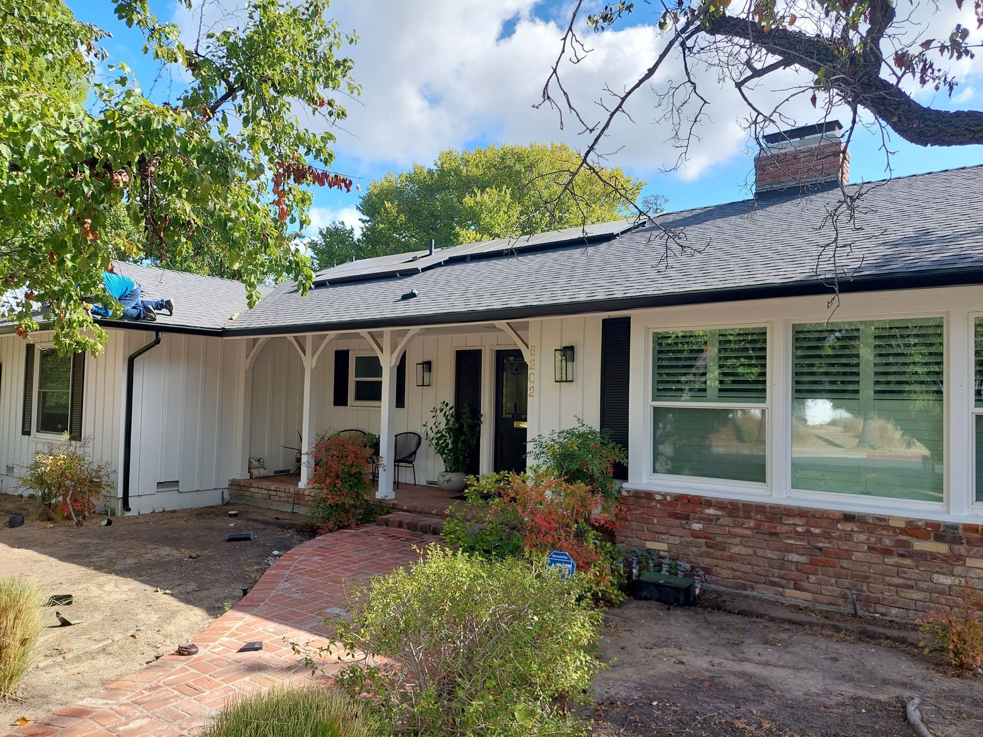 Single-story brick house with a wavy roof, white shutters, and a small front garden under trees.