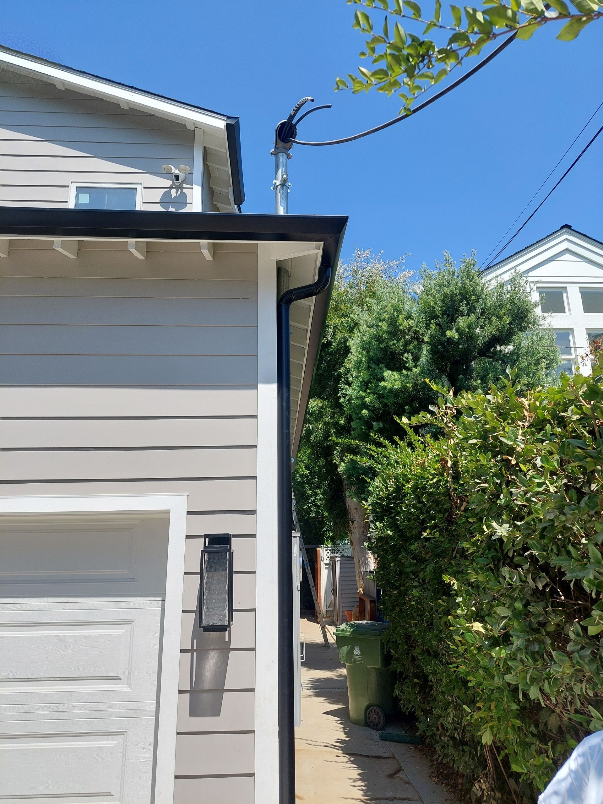 Side view of a light gray house and garage with a narrow passage, green hedge, and blue sky