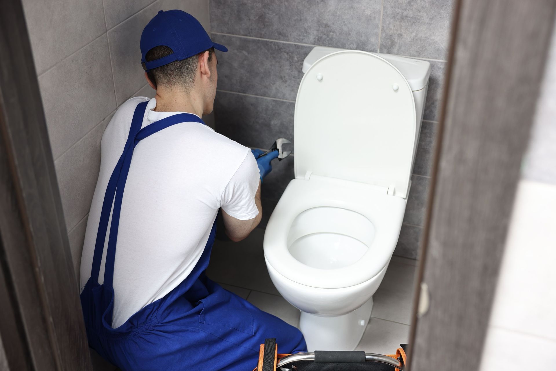 Plumber in blue overalls repairs a toilet in a tiled bathroom stall.
