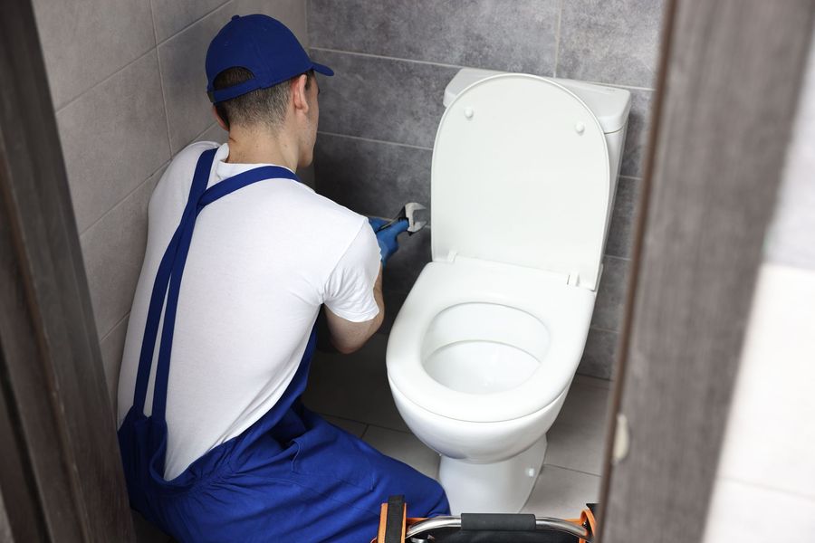 Plumber in blue overalls repairs a toilet in a tiled bathroom stall.