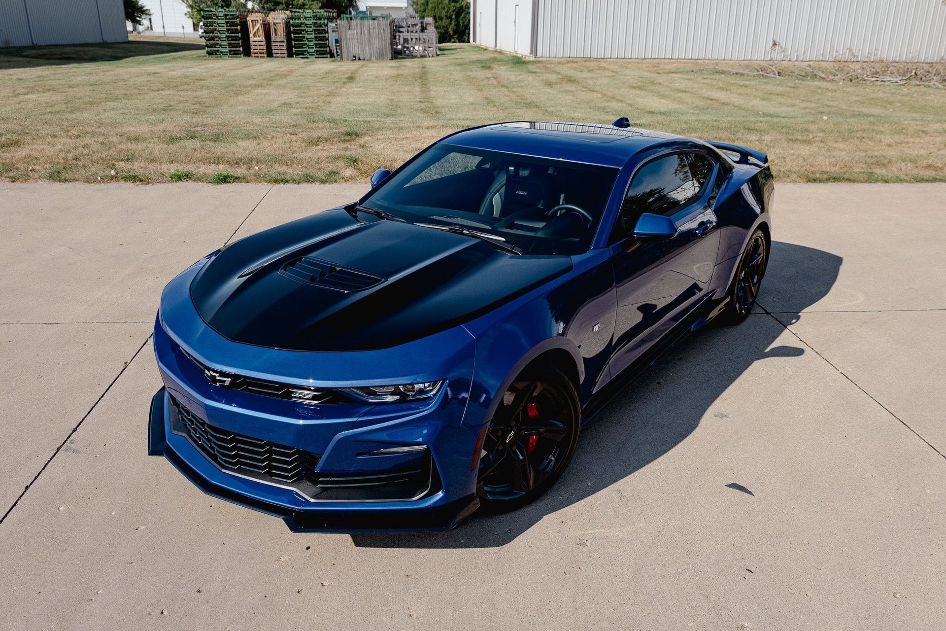 Blue sports car parked on a concrete lot near a chain-link fence and grassy field.