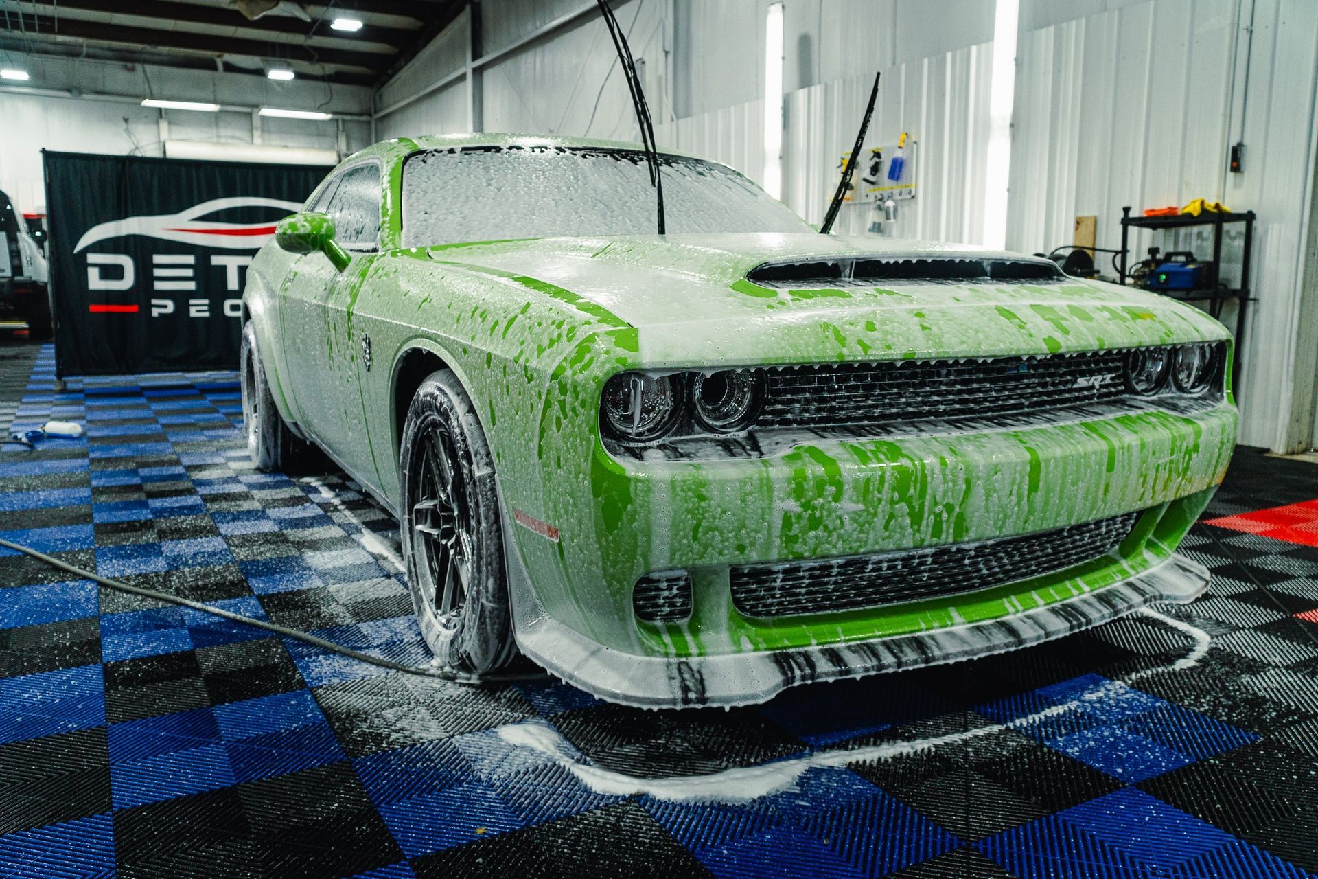 Green muscle car covered in soap foam inside an auto detailing shop with blue checkered floor mat