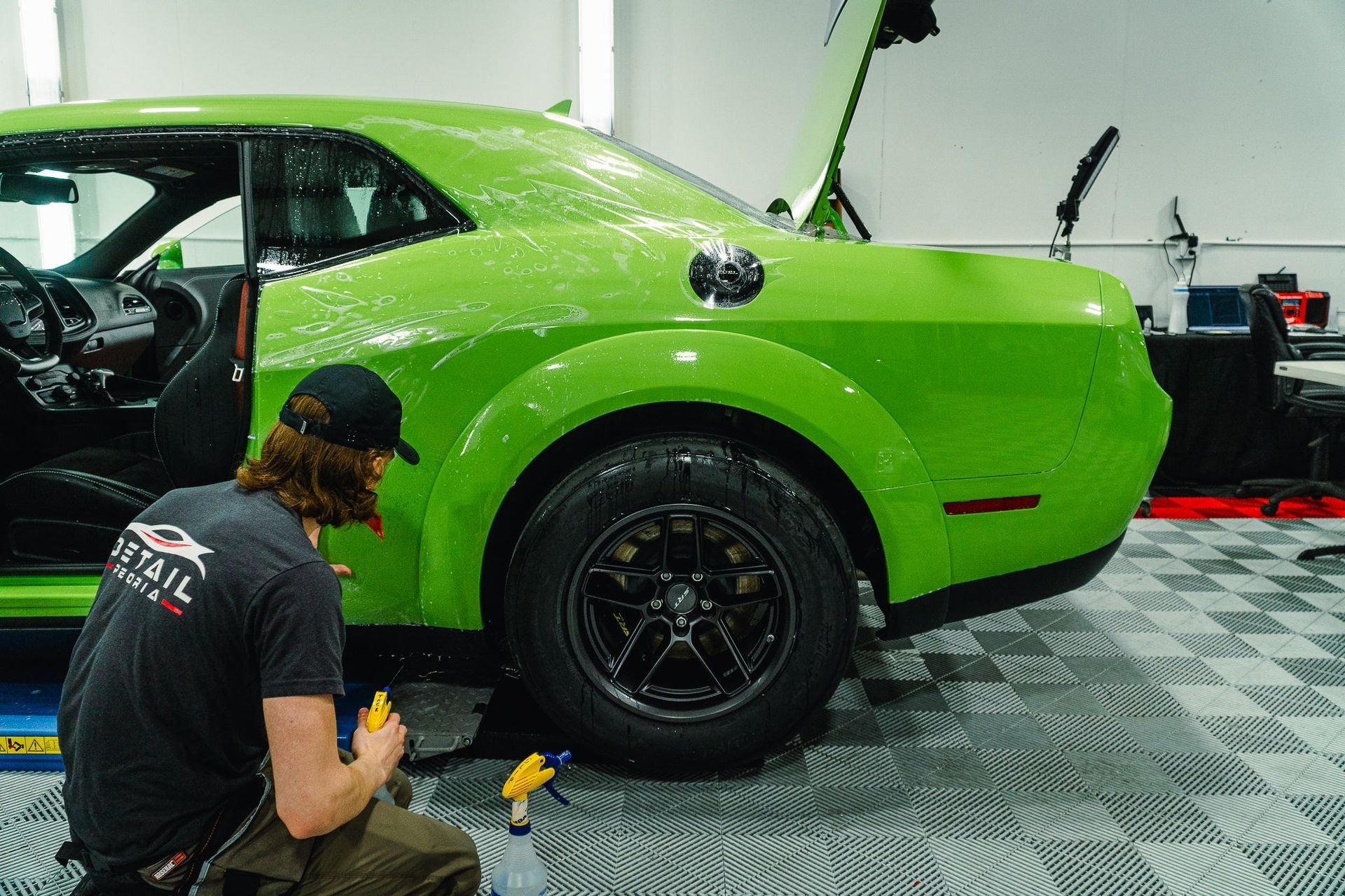 Green sports car in a garage with a mechanic crouching beside the rear wheel, working on it.
