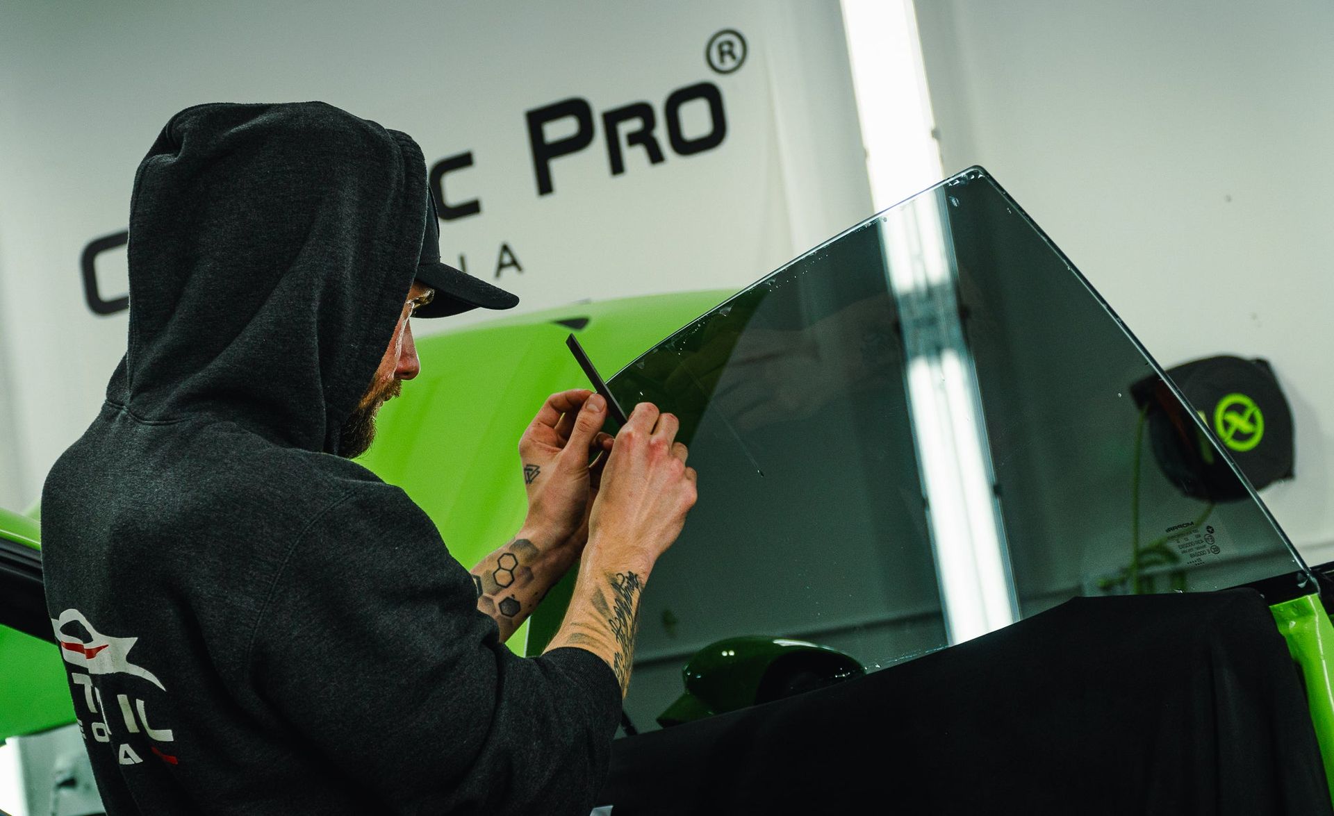 Person in a black hoodie applying vinyl to a glossy black vehicle wrap in a shop, with a green light reflection.