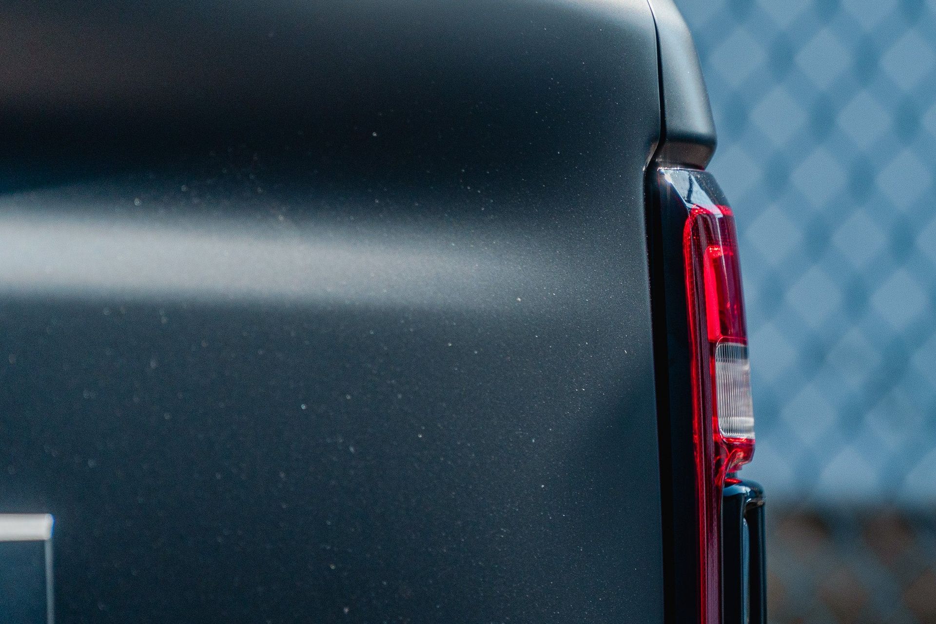 Close-up of a black vehicle’s rear corner with a red taillight, against a blurred blue background