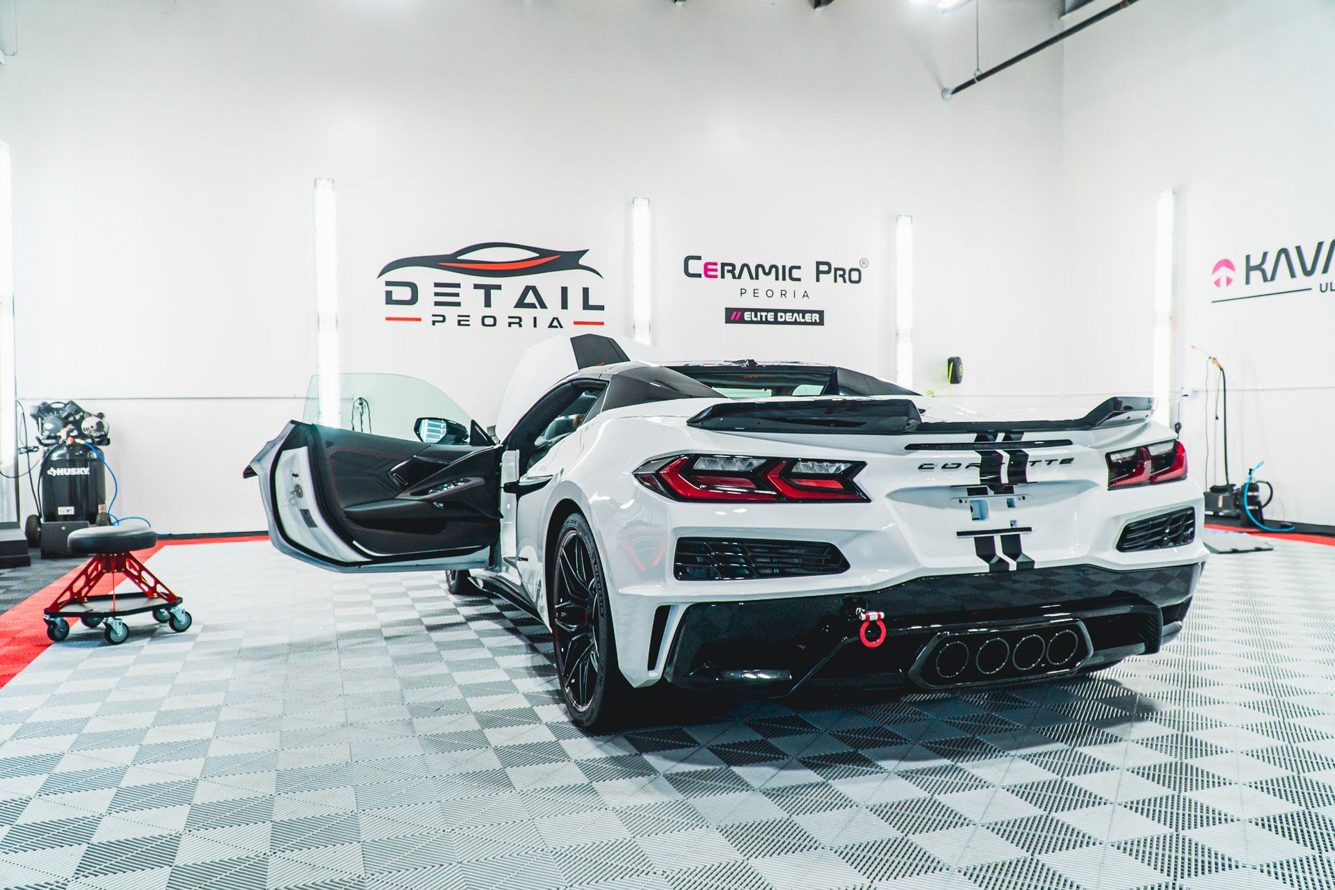 White sports car in a detailing shop, with doors open and a black roof spoiler on a checkered floor.