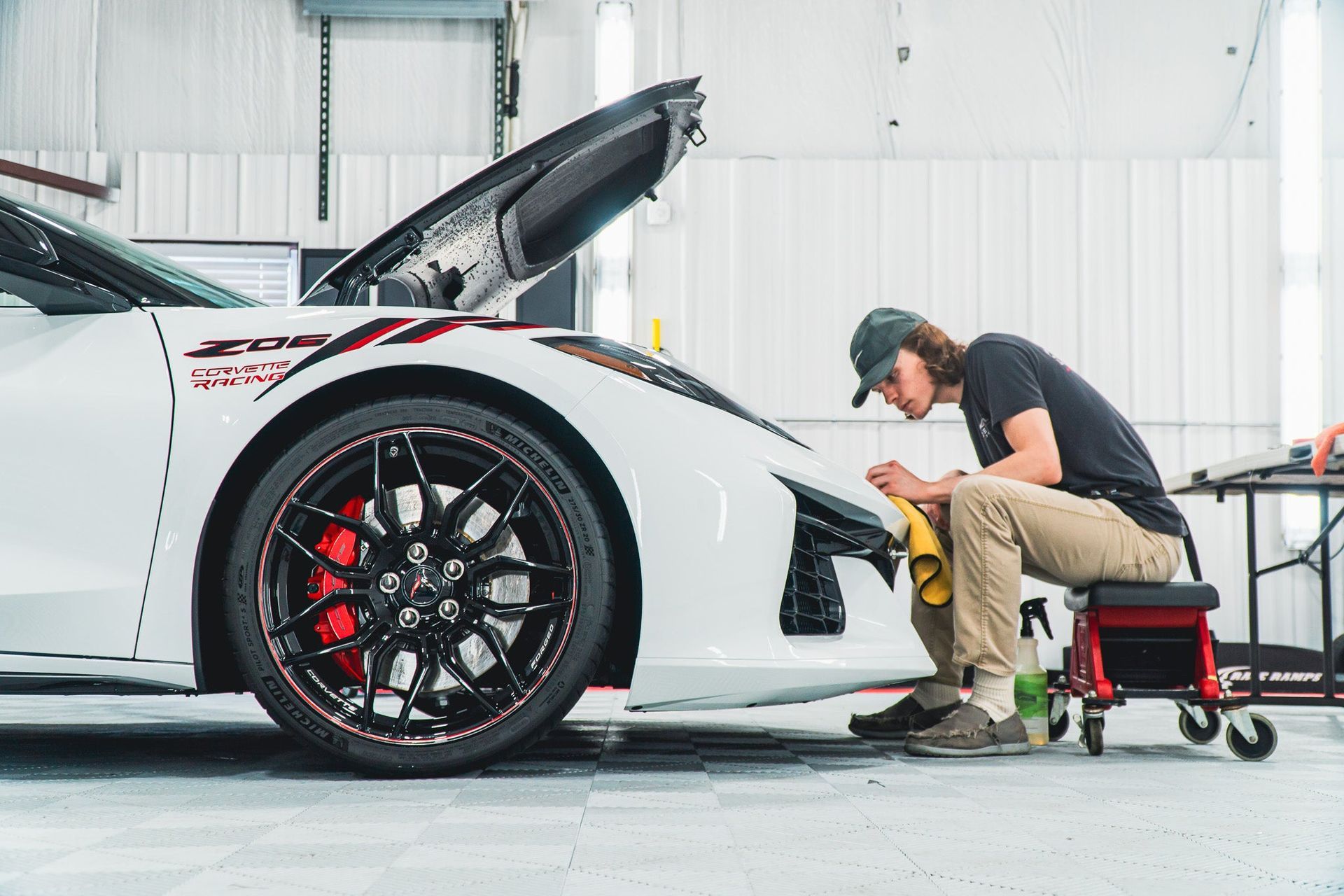 Mechanic crouches beside a white sports car with its hood open in a bright garage.