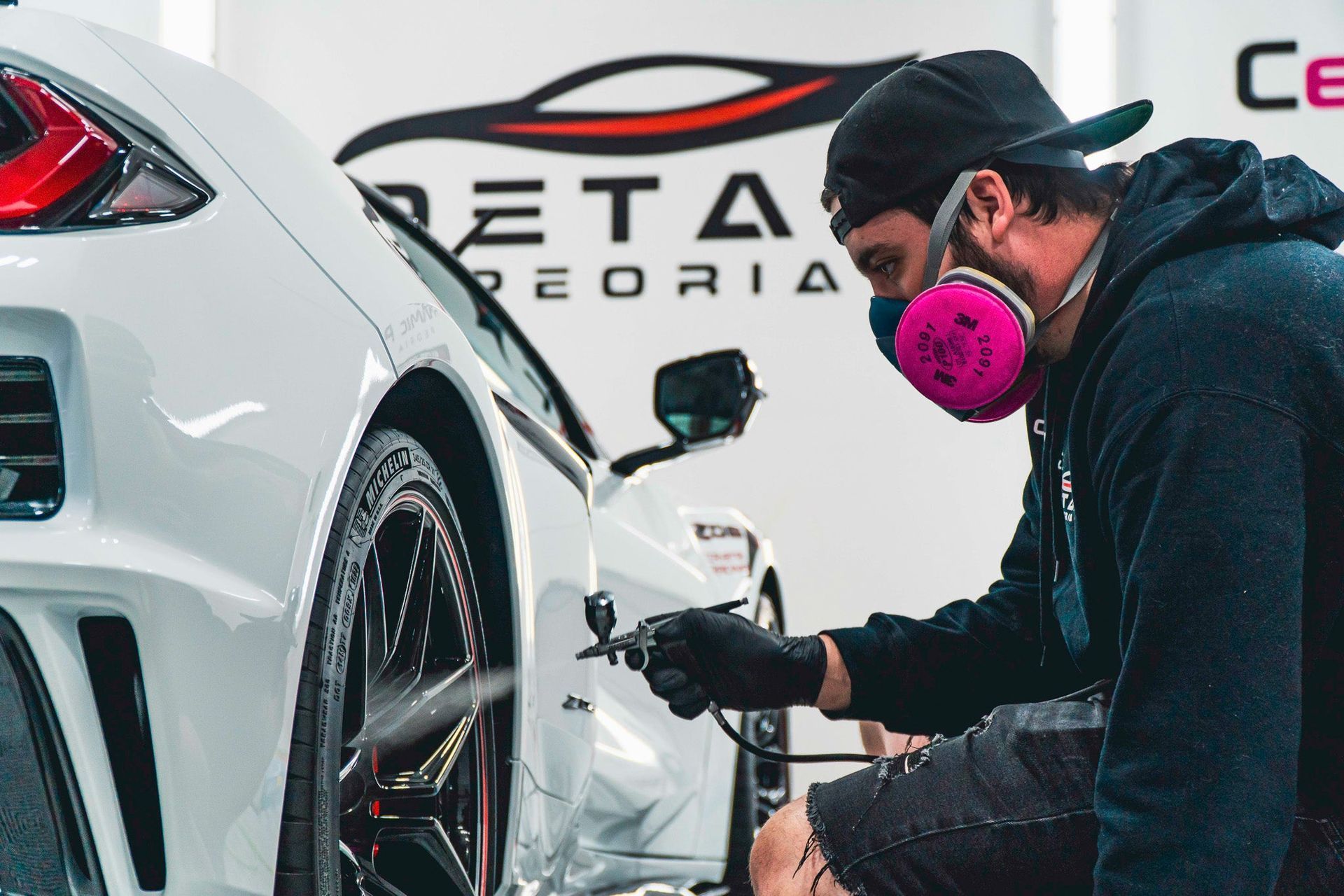 Detailing worker polishing a white car’s rear wheel in an auto shop, wearing a pink respirator mask