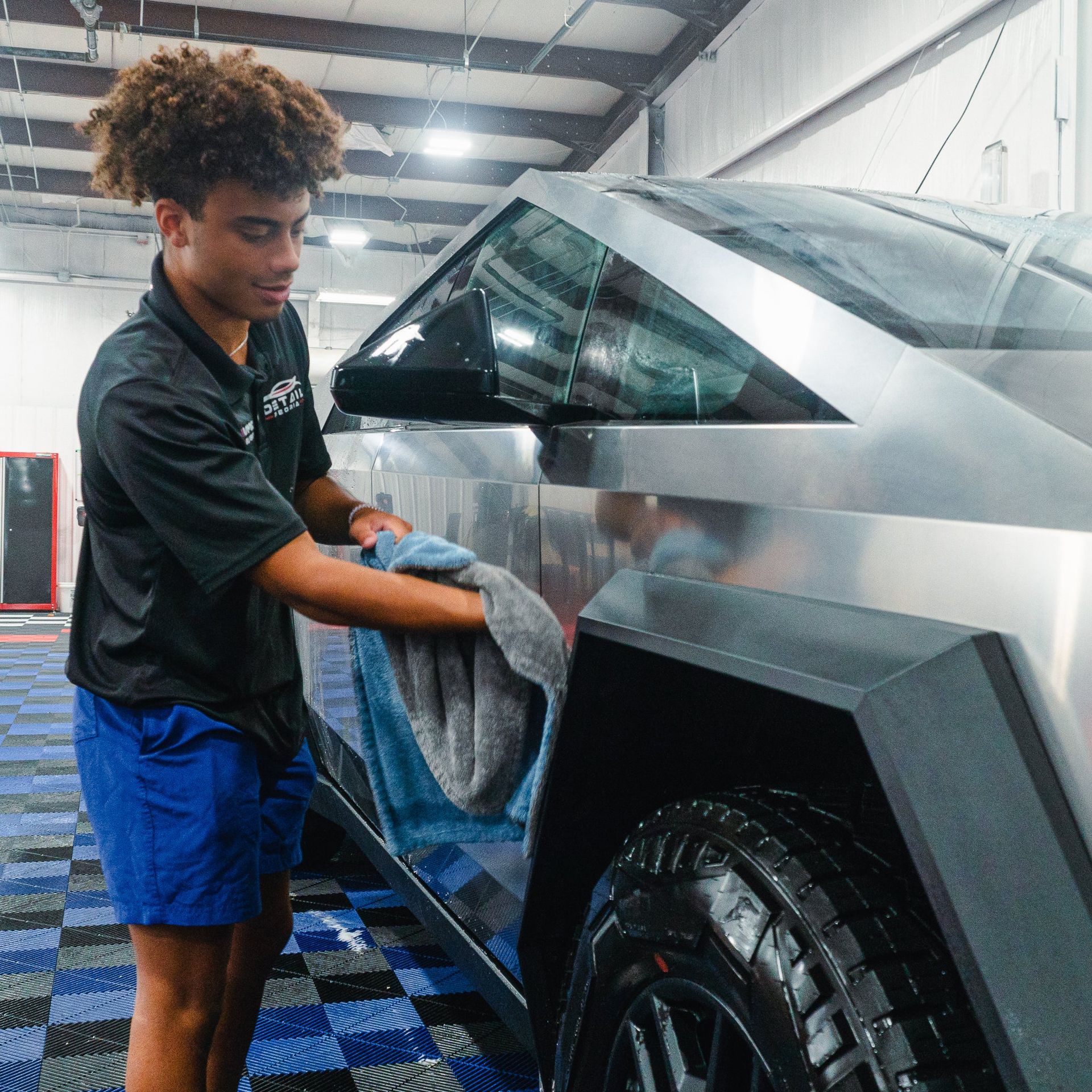 Person wiping a silver sports car in a garage with a blue checkered floor