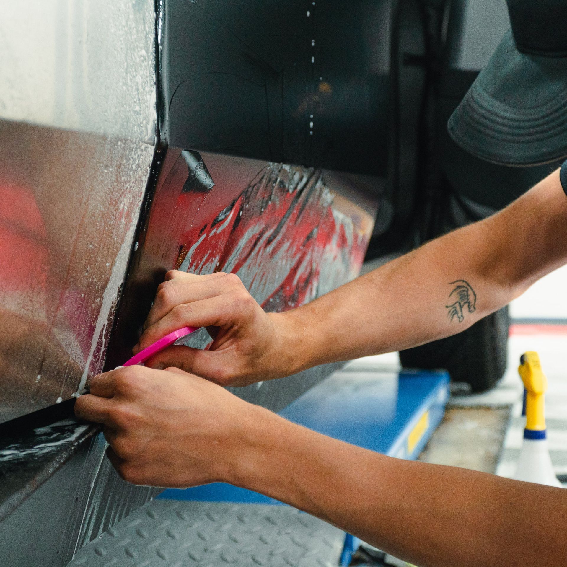 Hands peeling red vinyl wrap from a car panel with a pink tool