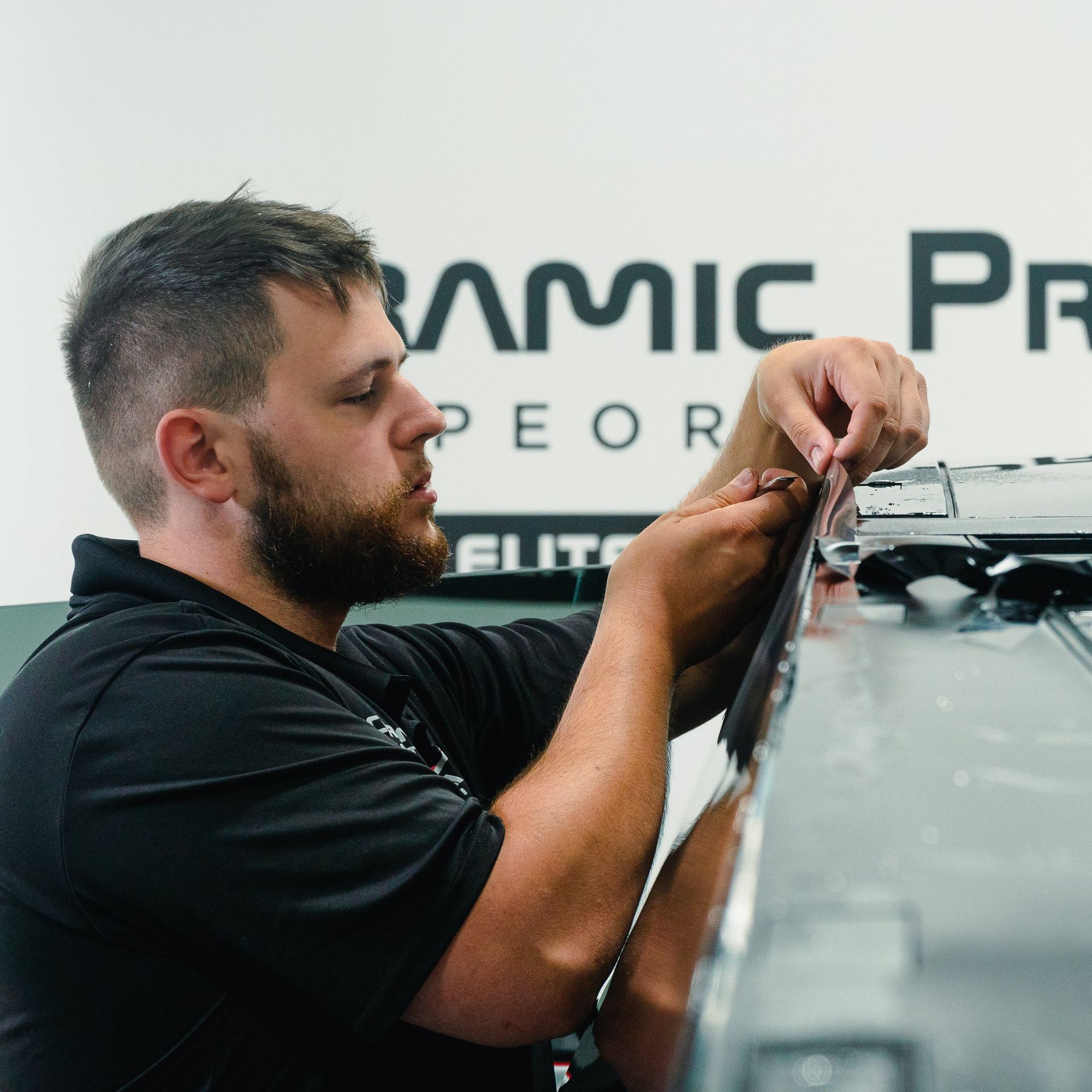 Technician working on car equipment indoors, adjusting wiring near a vehicle hood