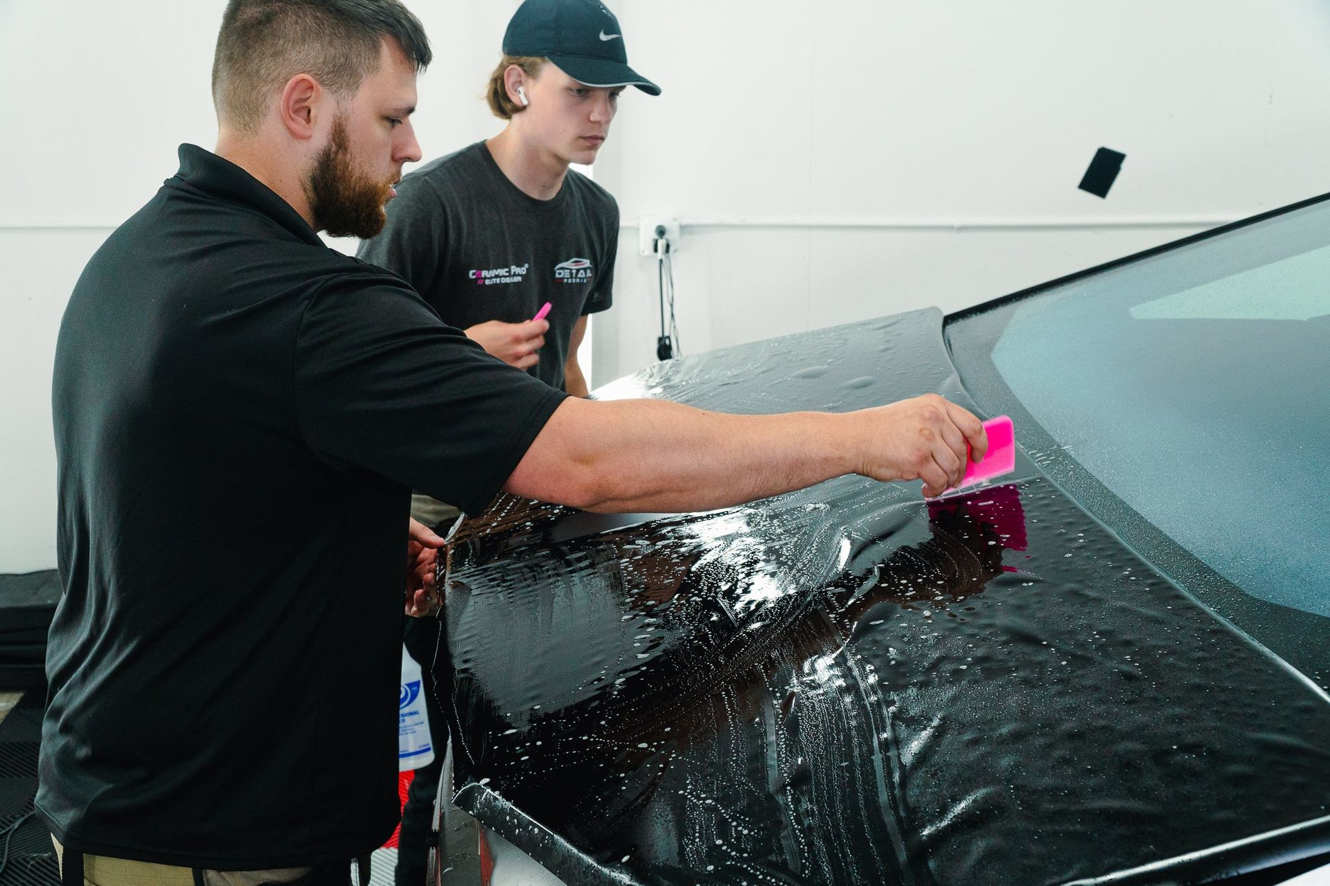 Two people wash a black car windshield with a pink sponge.