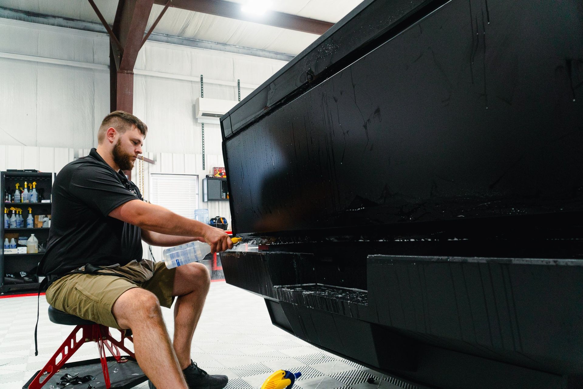 Man painting a black truck bed in a garage workshop, wearing gloves and sitting on a stool