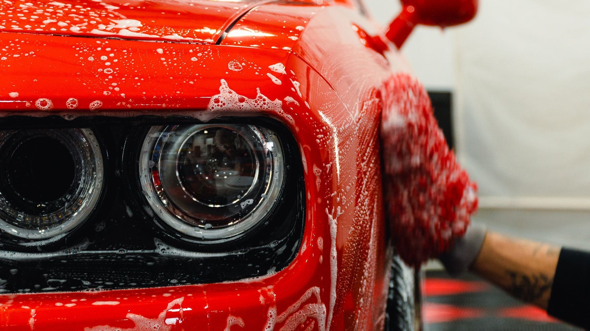 Close-up of a red car being washed, with water and soap on the hood and a person’s arm scrubbing