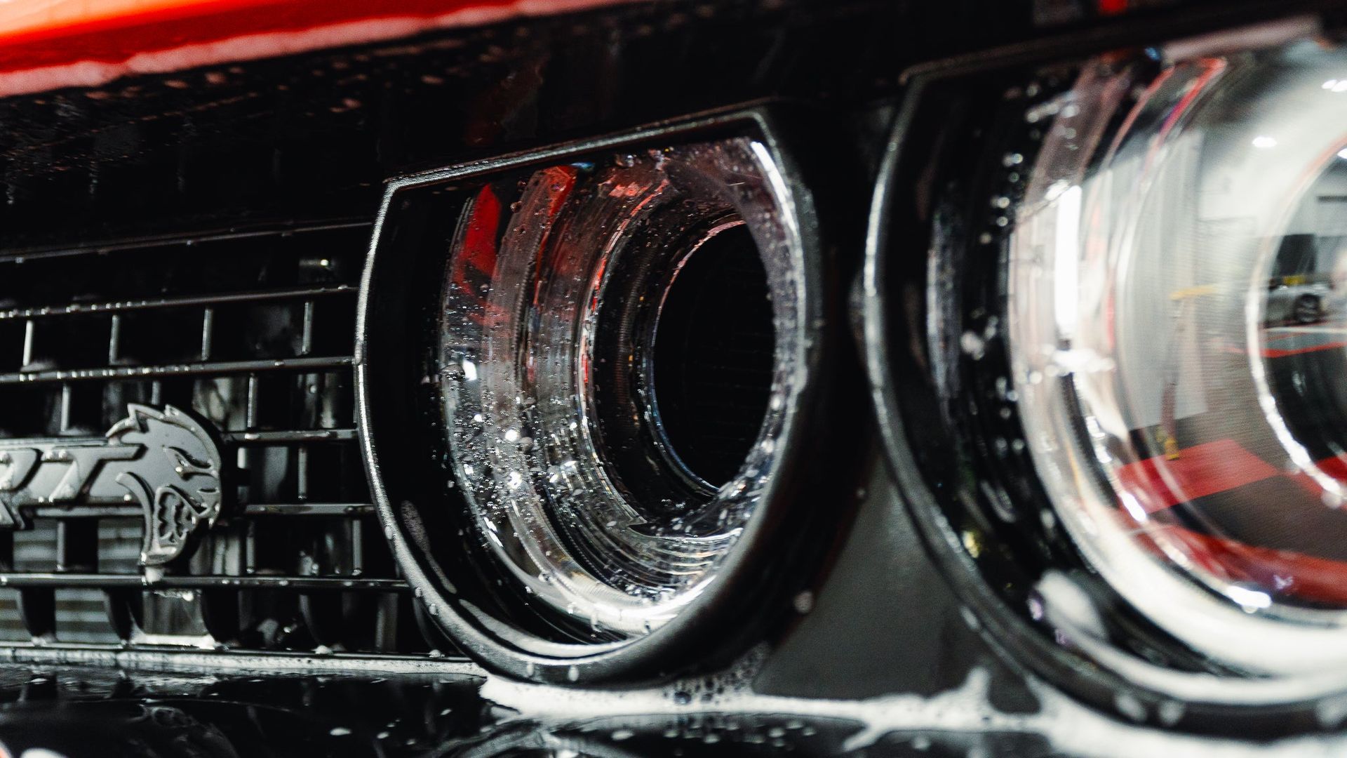 Close-up of a red sports car grille with two chrome headlights splashed with water