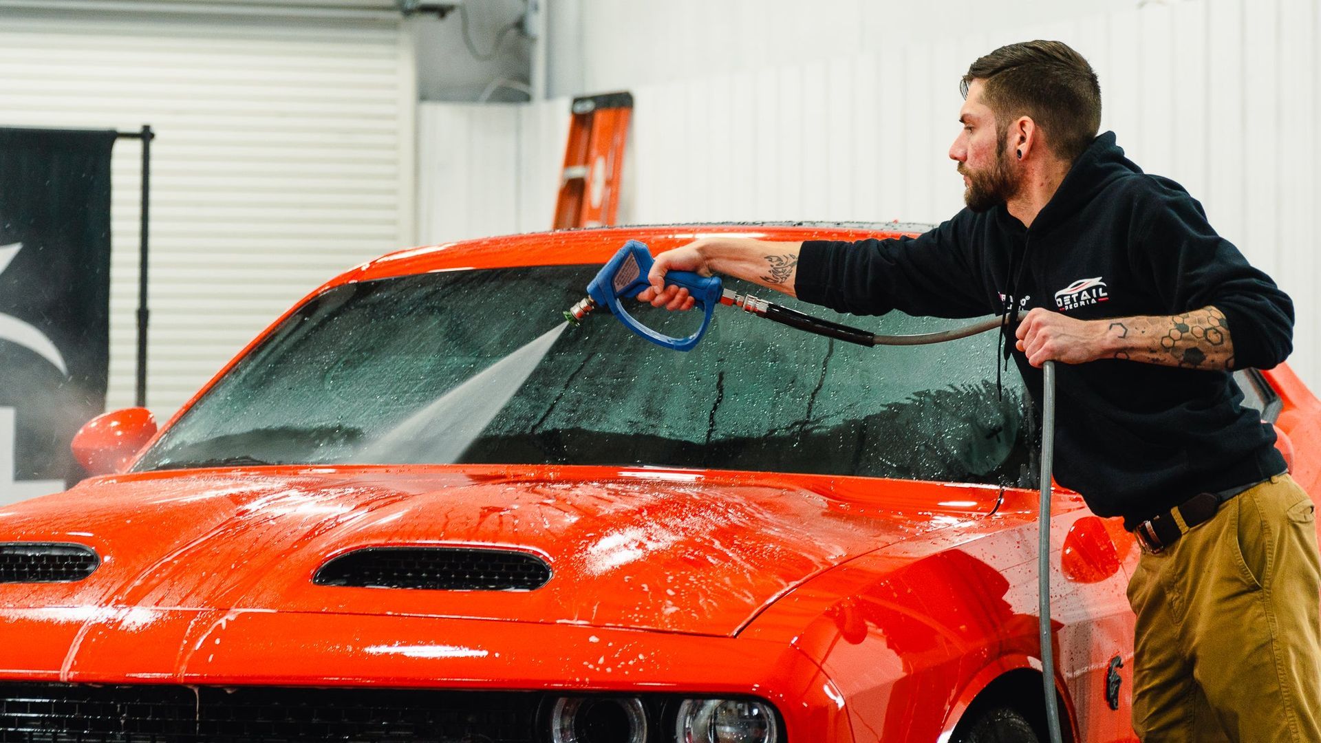 Man washing a red sports car in a garage, spraying foam across the windshield