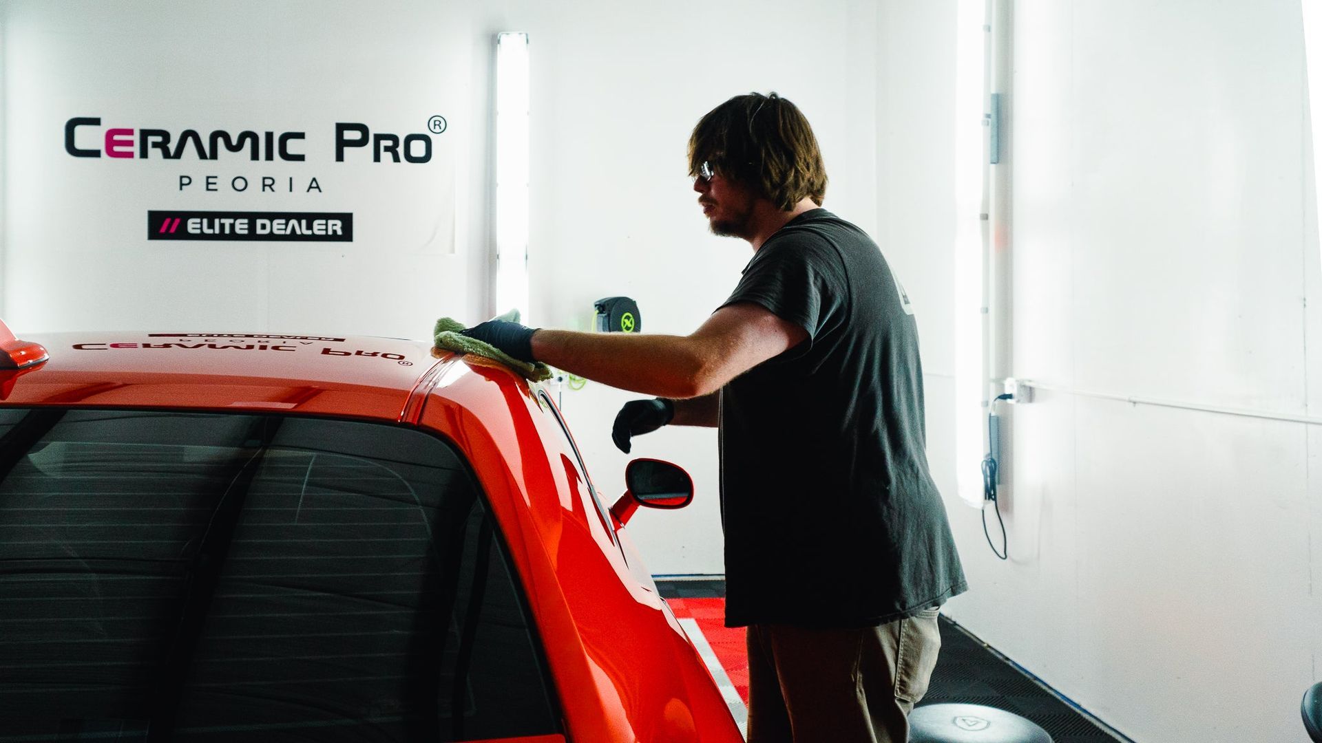 Detailing technician polishing a red car beside a Ceramic Pro sign in a bright shop