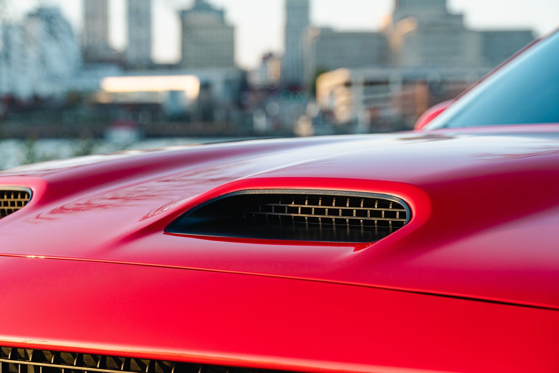Red sports car hood with air vents, city skyline blurred in the background
