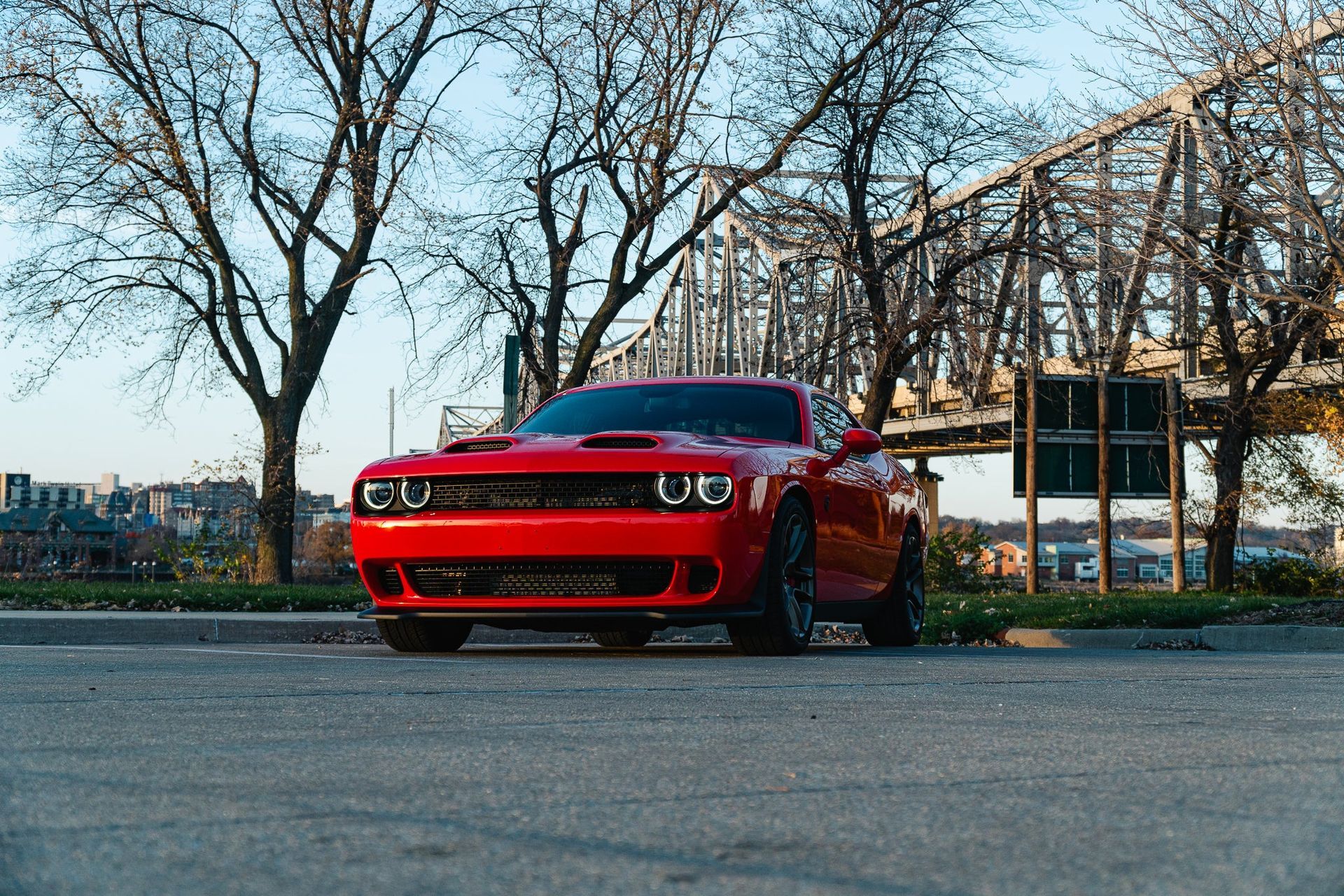 Red sports car parked on pavement near leafless trees and a bridge at dusk