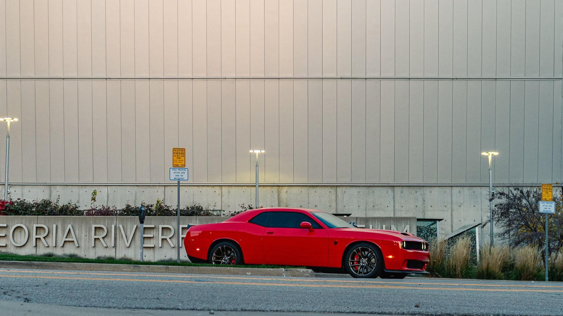 Red sports car parked beside a stone wall with “Peoria River” lettering at dusk