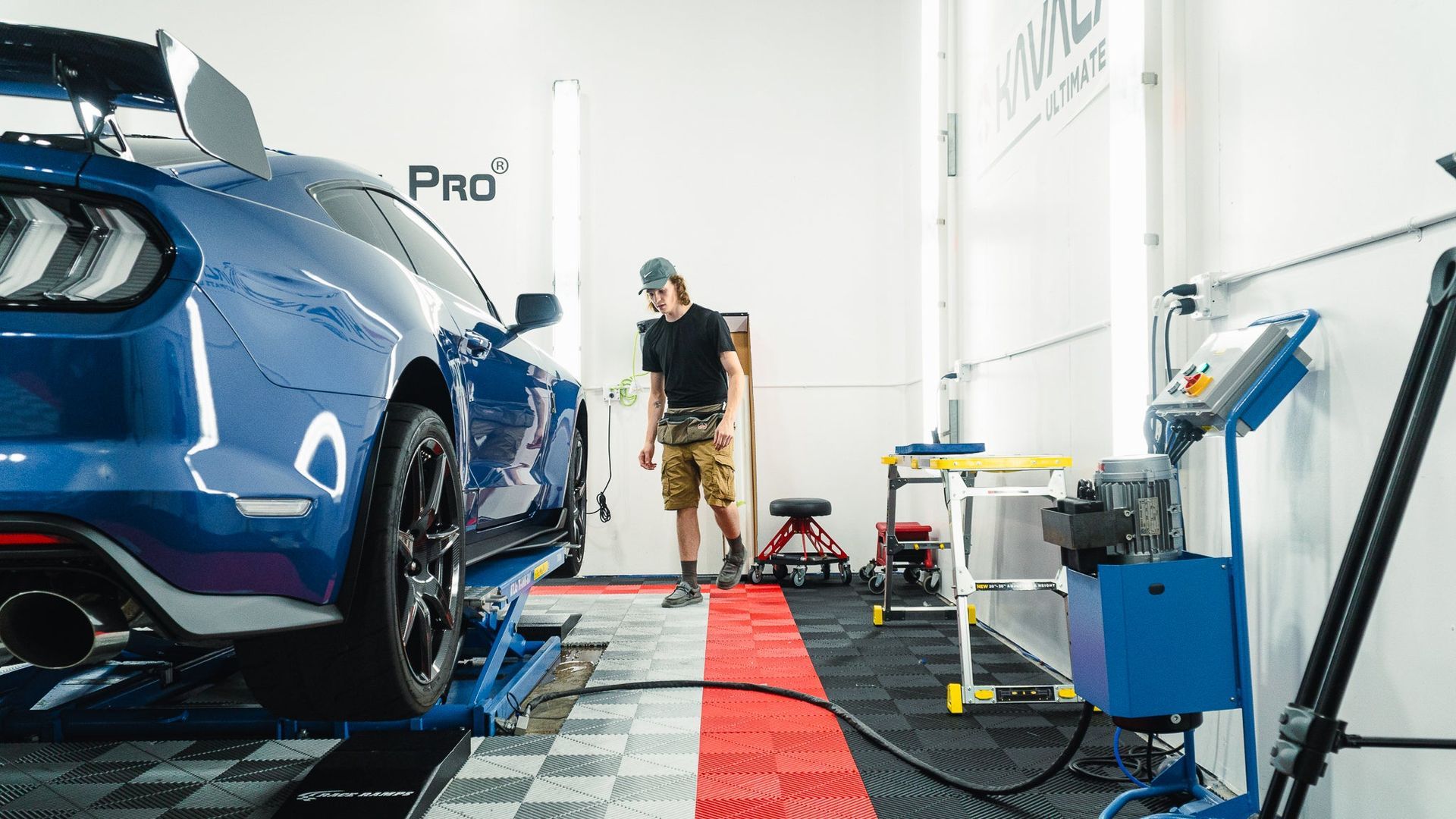 Blue car in an auto shop, with a mechanic standing beside tools and equipment on a red floor mat.