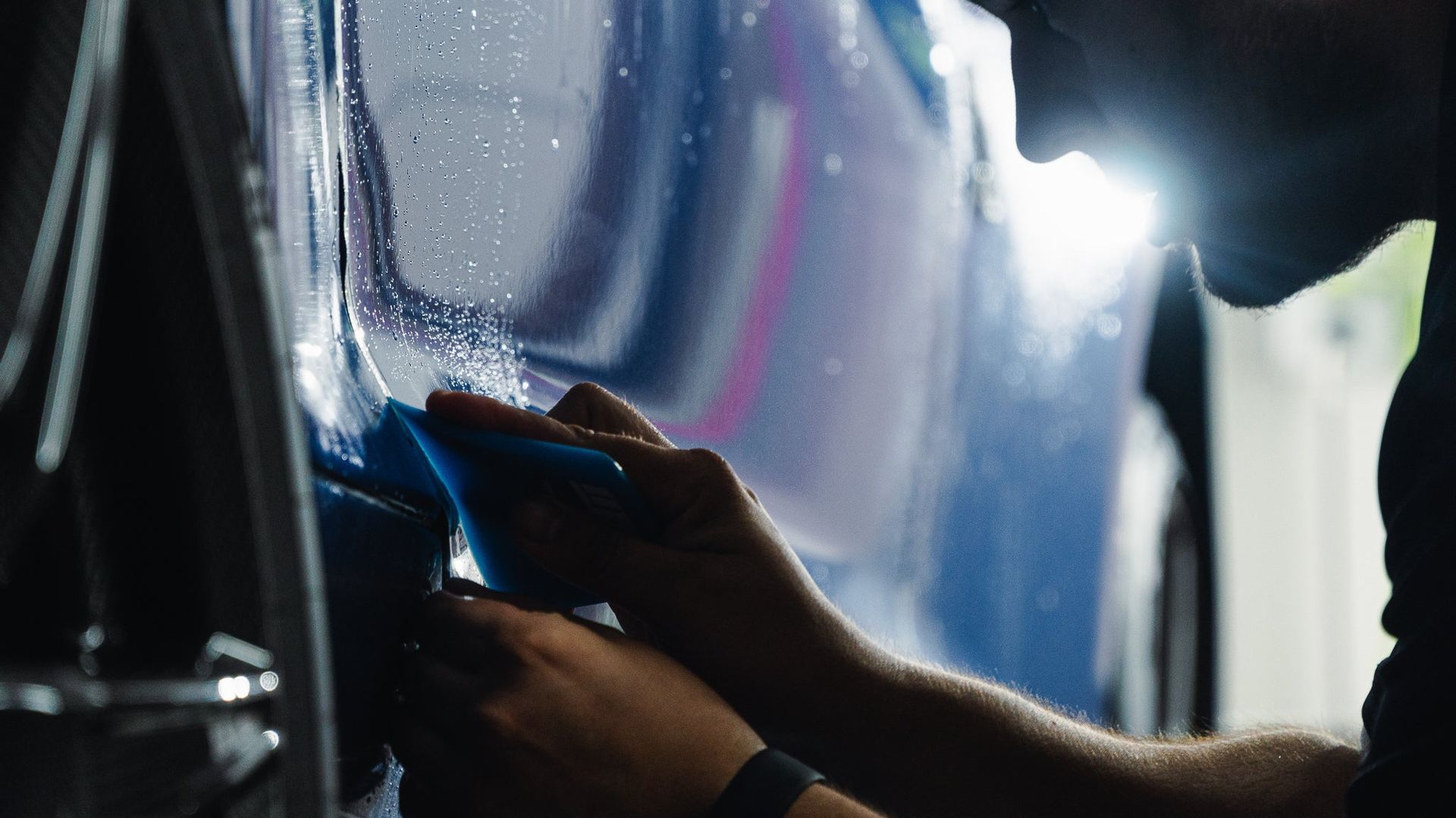 Hands polishing a glossy blue car under bright lights in a dark garage