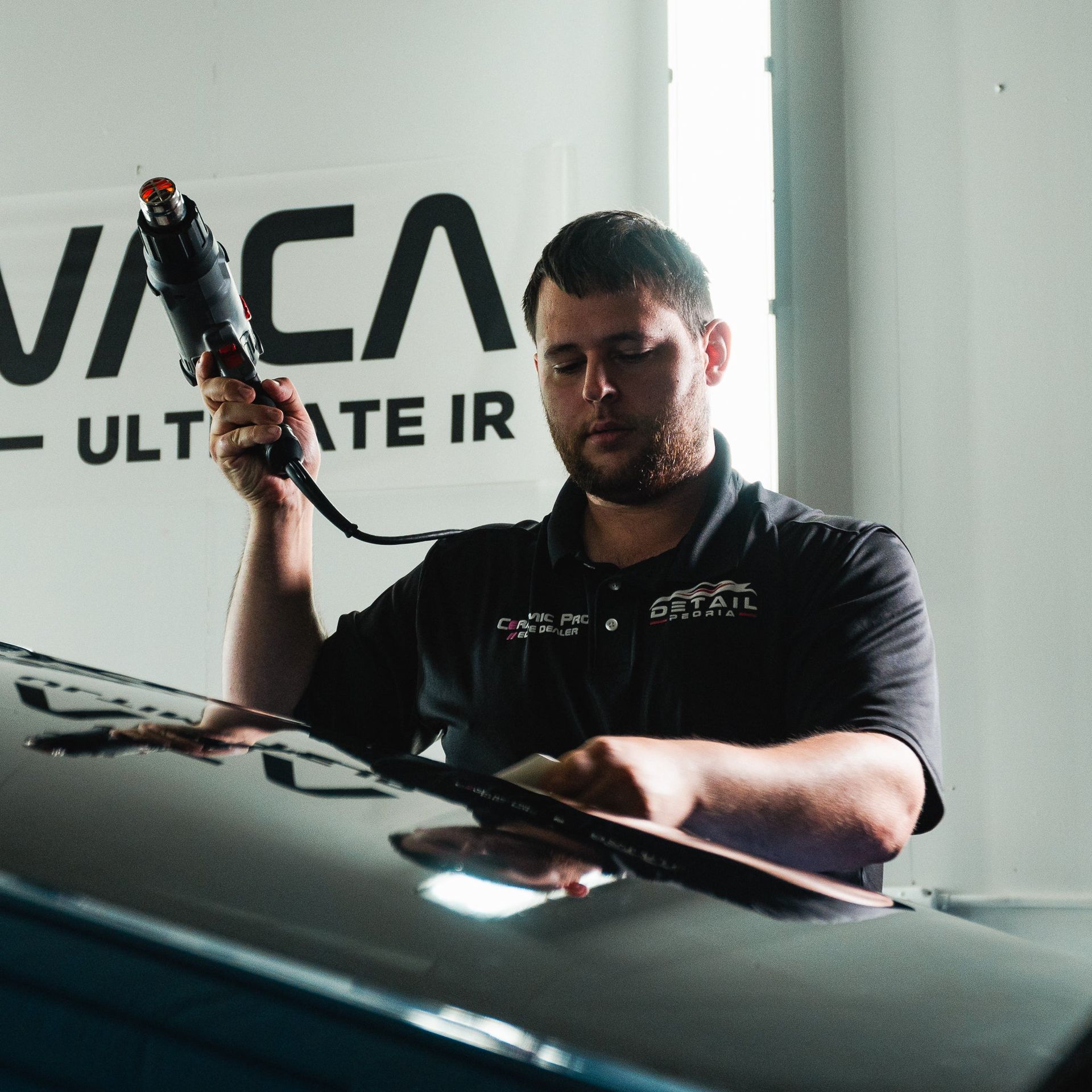 Man polishing a black car with a tool in a garage, with a VCA banner behind him