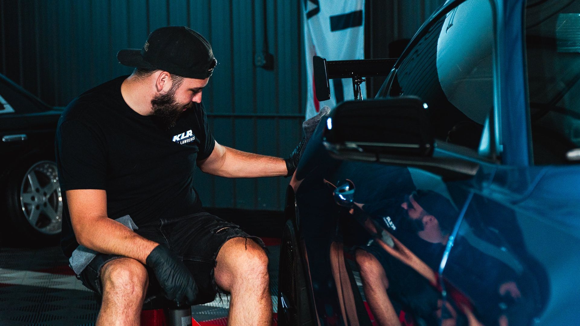 Man inspecting a blue car’s interior in a dim garage, with the hood open.