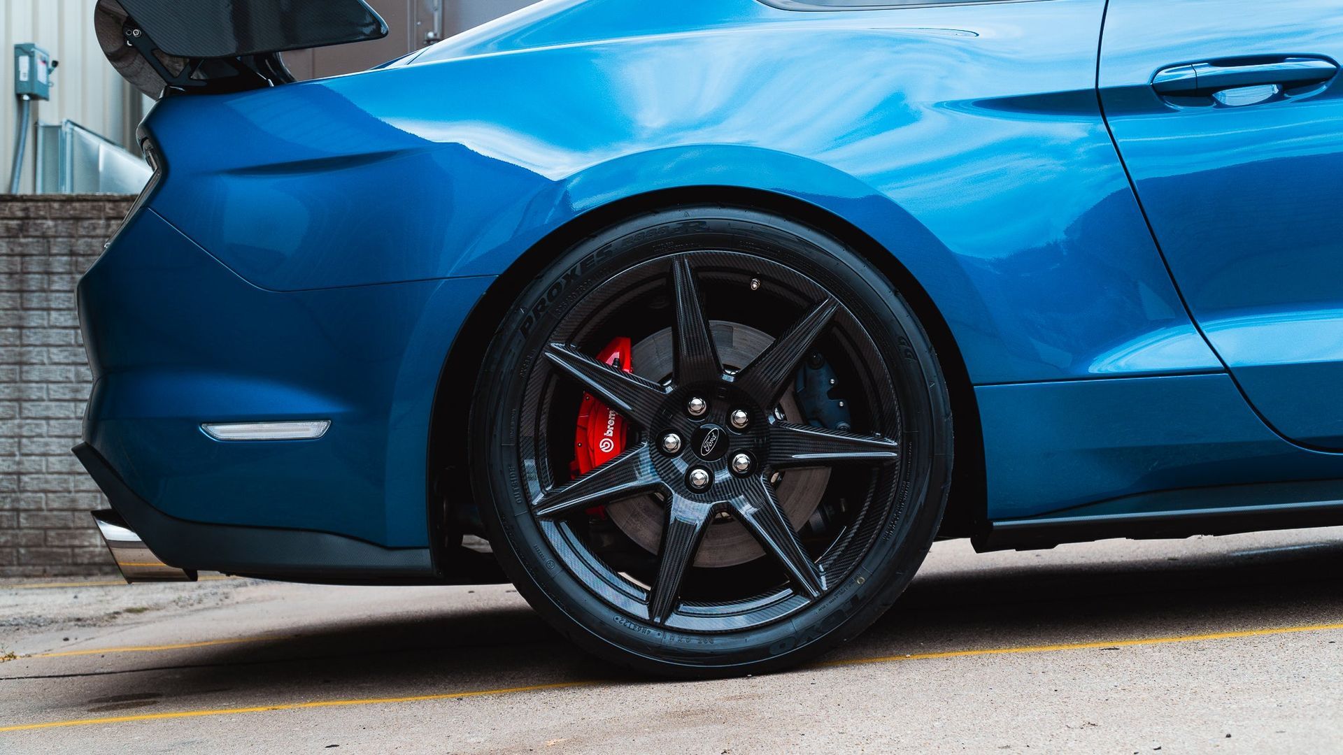 Blue sports car rear wheel and side panel with black rims and red brake caliper, parked on pavement