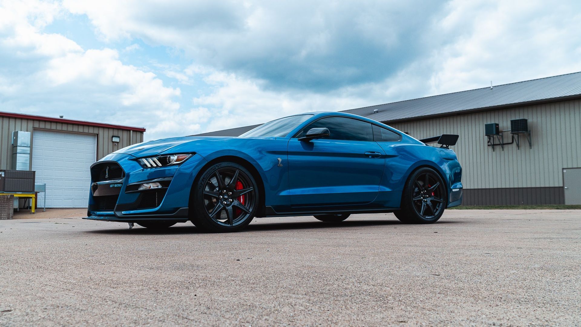 Blue sports car parked on pavement outside an industrial building under a cloudy sky