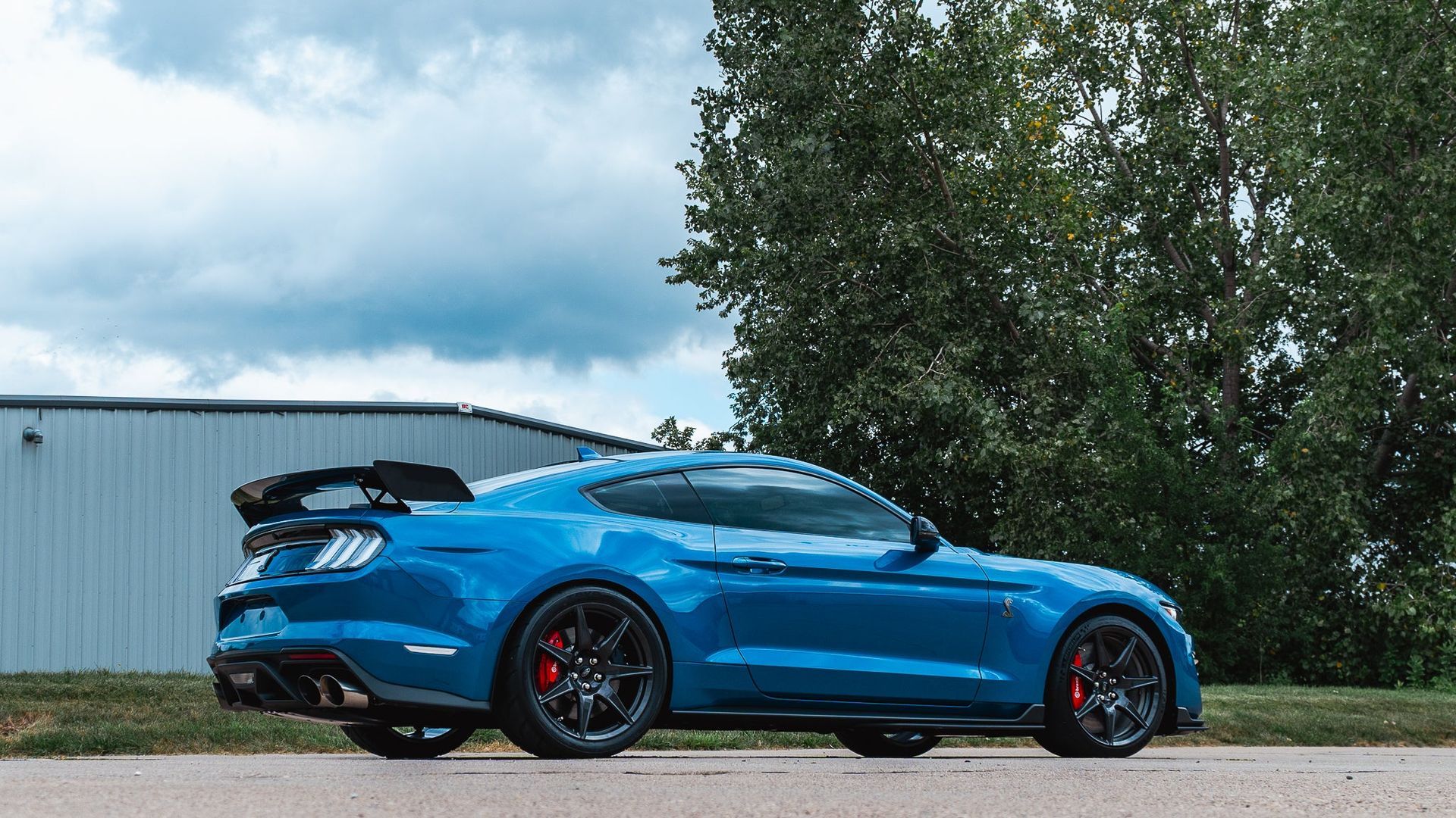 Blue sports car parked outdoors beside a metal building under cloudy skies.