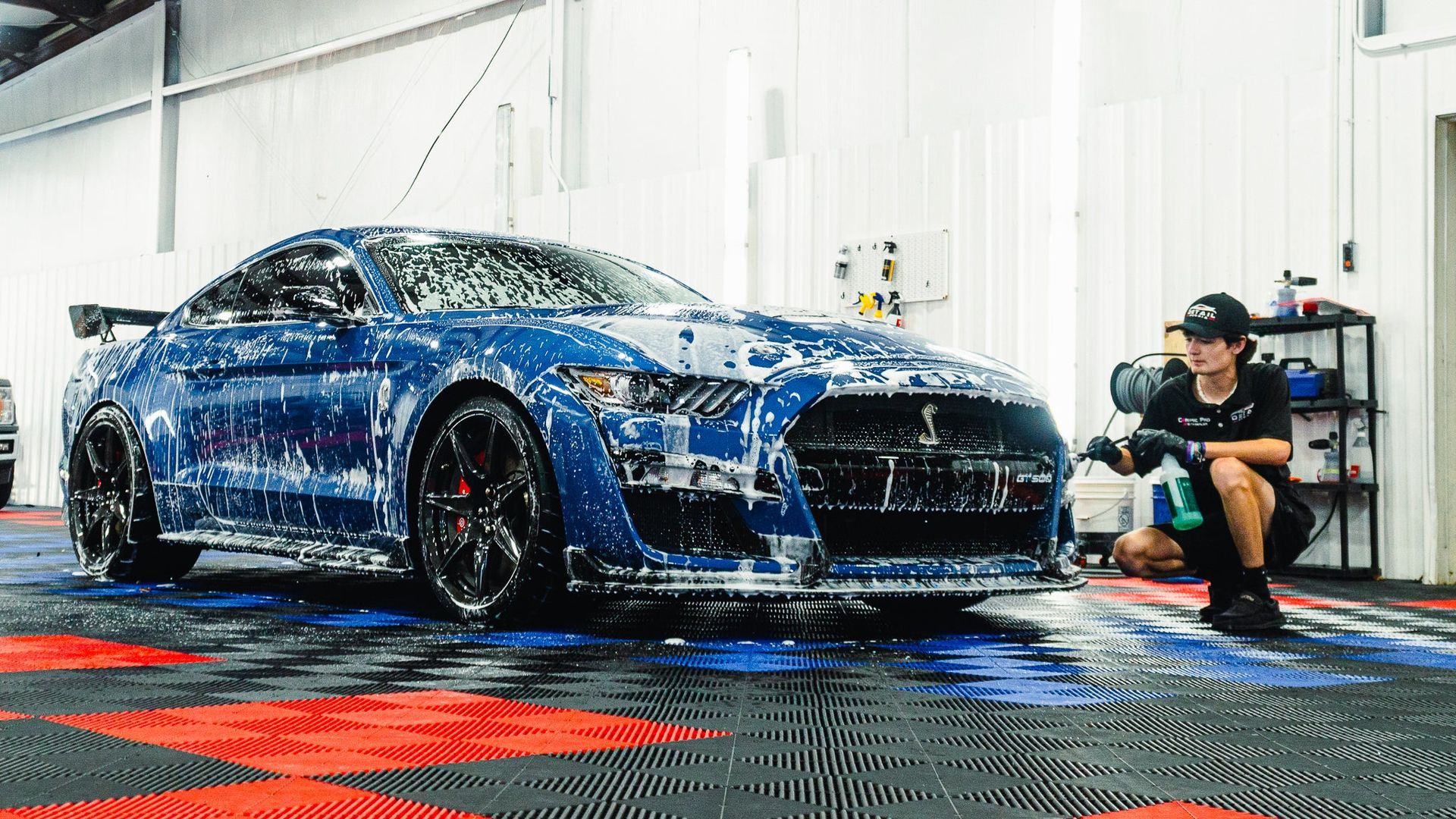 Blue sports car being detailed in a garage, with a worker polishing its front wheel.