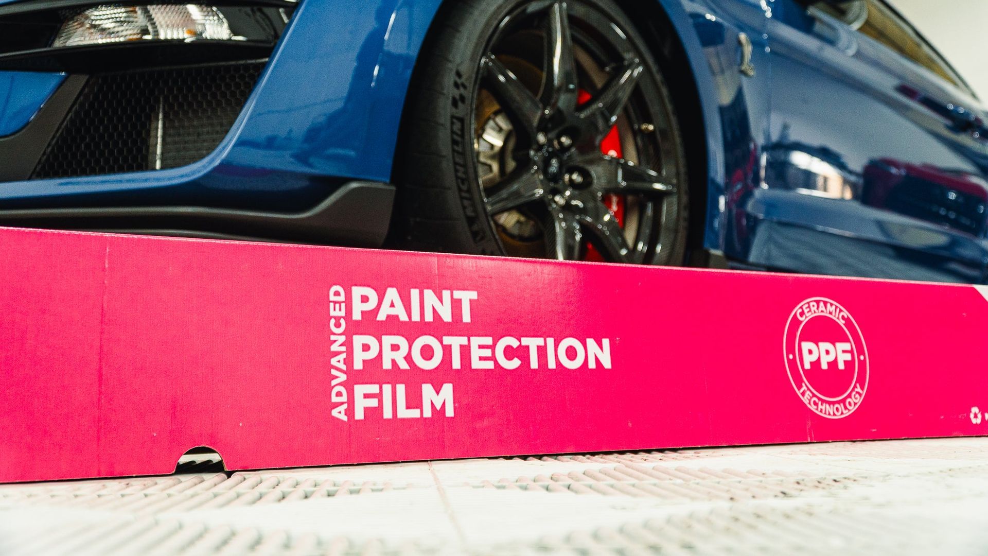 Blue sports car behind a pink “Paint Protection Film” display board