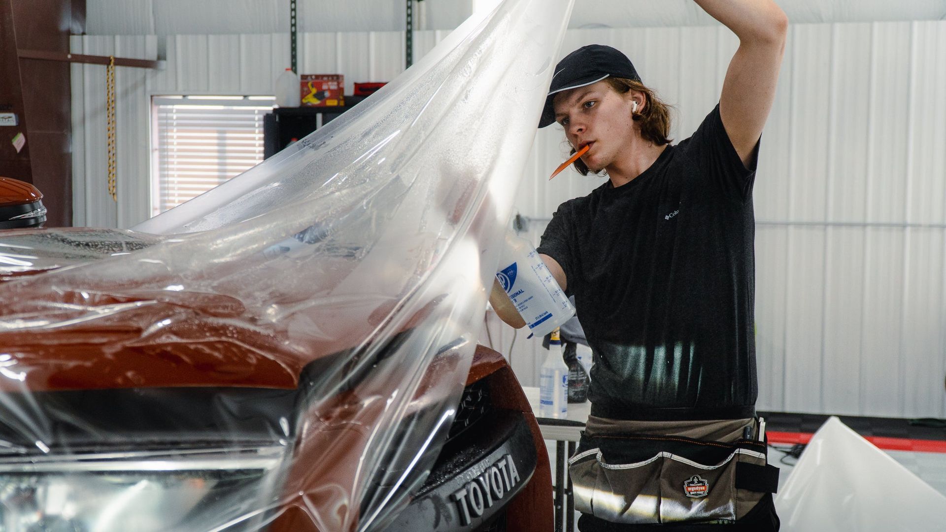 Person wrapping a red car in clear protective film inside a garage-like workshop.