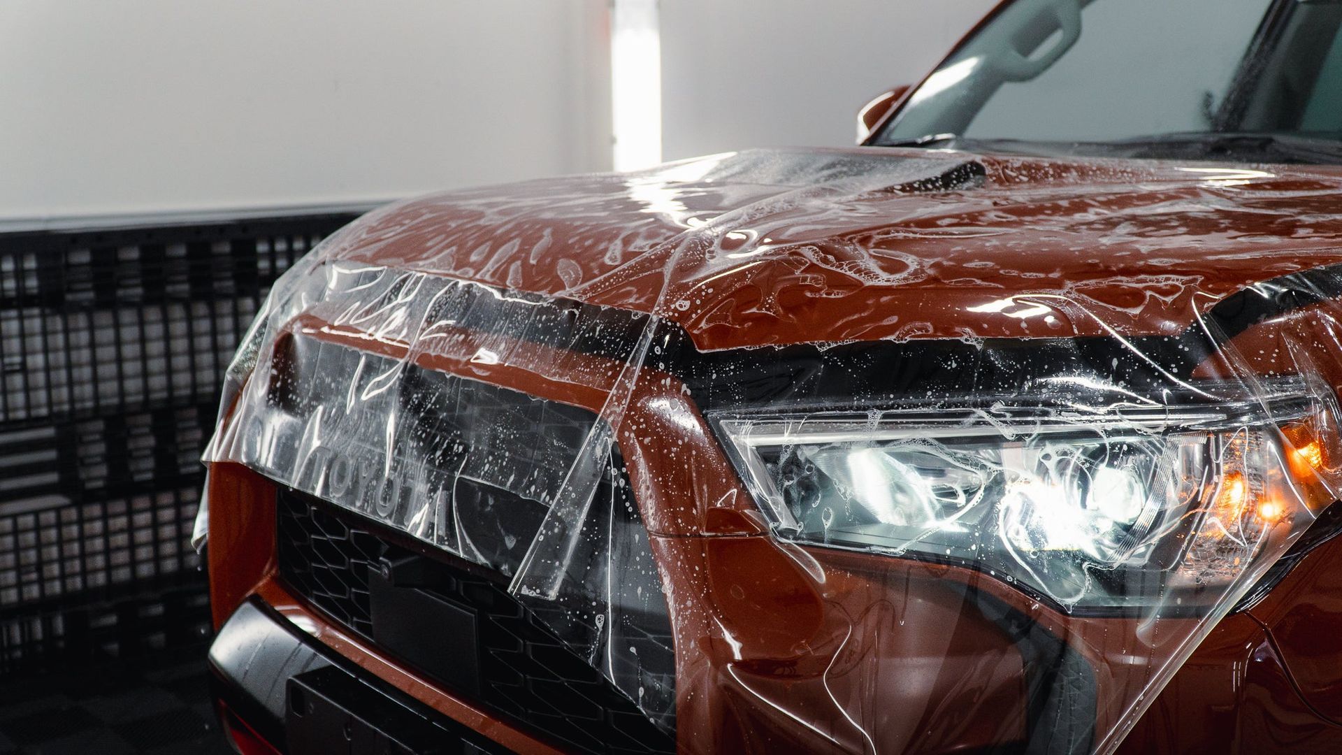 Red car covered in protective plastic, viewed from the front in a garage setting