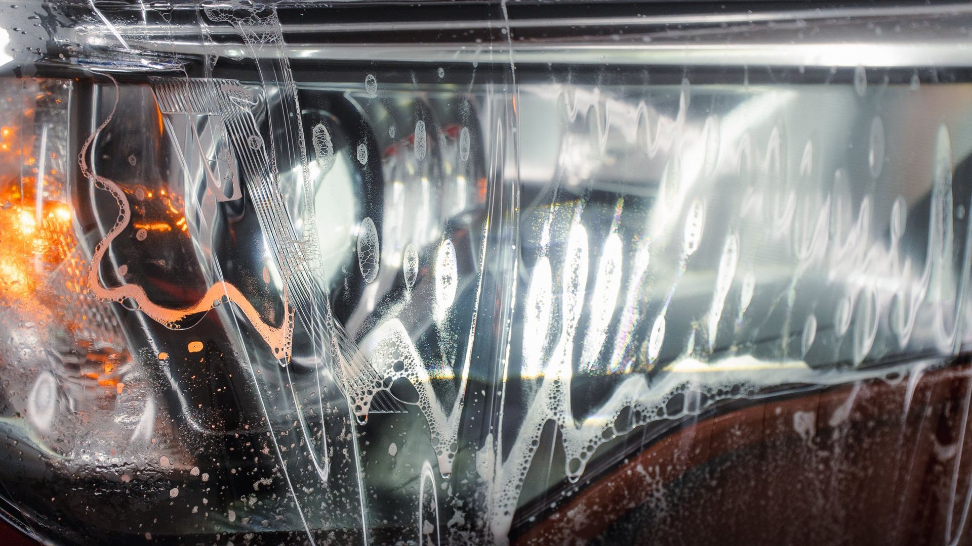 Car headlight shining through rain-streaked windshield, with orange reflections on the glass