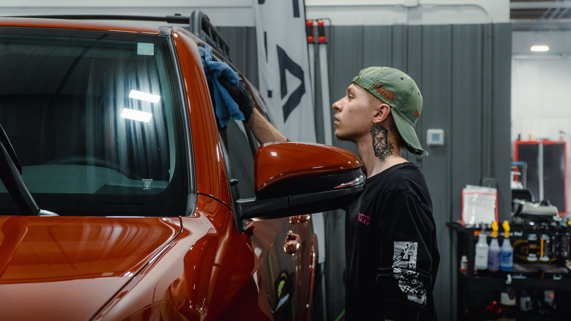 Person polishing an orange truck in a workshop, wearing a green cap and black shirt